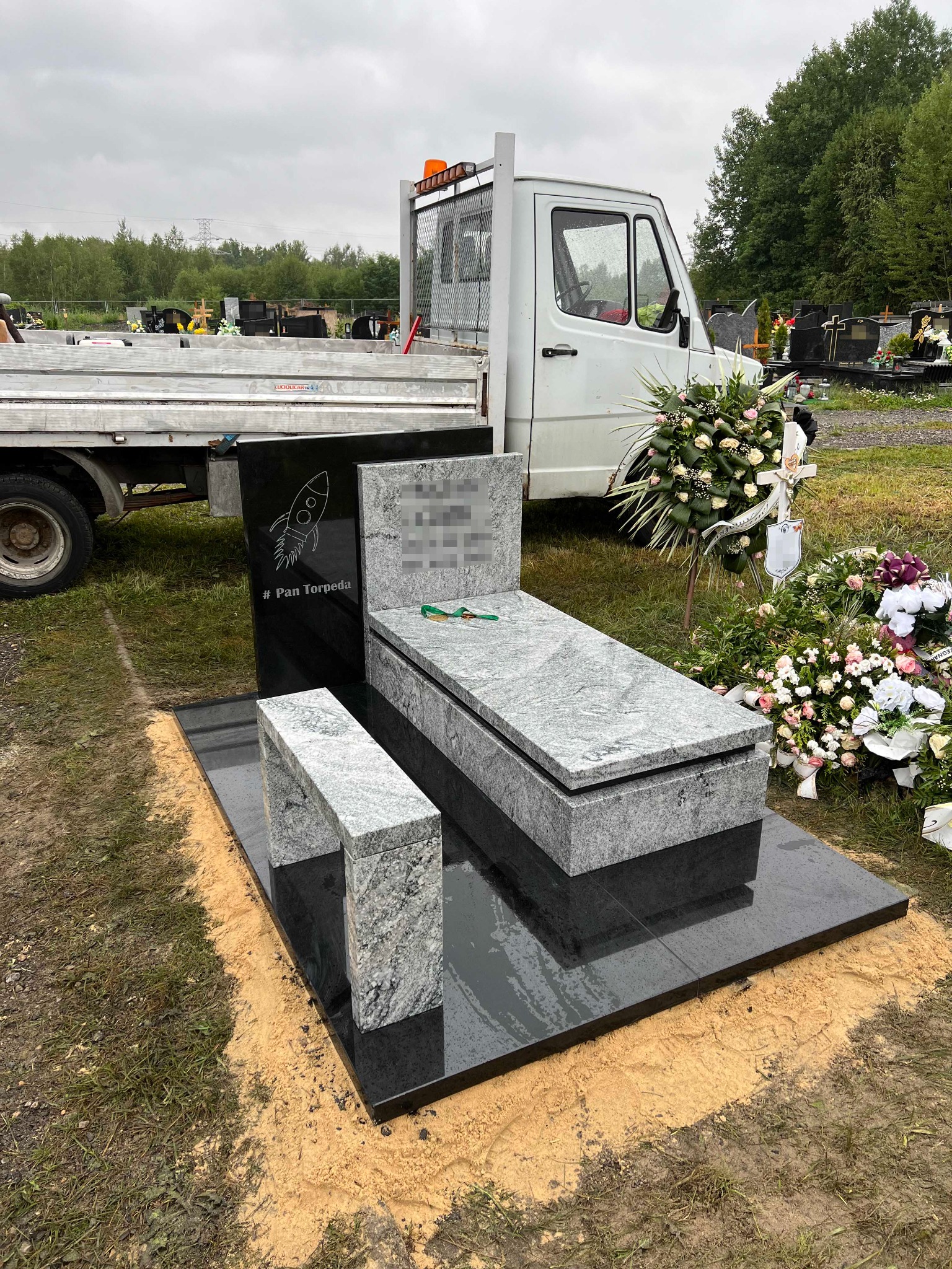 Czarny nagrobek z jasnoszarym nakryciem i ławką, ozdobiony grawerem rakiety i napisem '# Pan Torpeda', ustawiony na cmentarzu, obok dekoracje kwiatowe i samochód dostawczy.