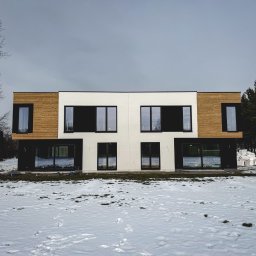 Karsbud - Nowoczesny, dwukondygnacyjny dom z elewacją łączącą biały tynk i drewniane panele. Czarne ramy okienne i drzwi. Śnieg na trawniku. Pochmurne niebo.