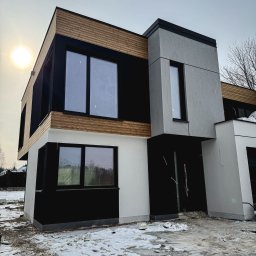 Karsbud - Nowoczesny dom z elewacją w odcieniach bieli, szarości i drewna. Widoczne okna i fragmenty instalacji. Na ziemi śnieg. Słońce w tle.