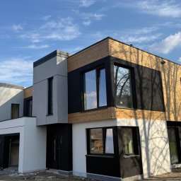 Karsbud - Nowoczesny dom z elewacją w odcieniach szarości, bieli i drewna, z dużymi oknami i płaskim dachem, na tle błękitnego nieba z chmurami. Widoczne prace wykończeniowe.