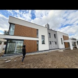 Karsbud - Nowoczesny dom z elewacją w odcieniach bieli, szarości i drewna. Widoczna osoba w pobliżu budynku. Uwagę przykuwają duże okna i minimalistyczny design.