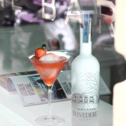 BRUNON FIDRYCH - Koktajl w kieliszku martini z truskawką i lodem, obok butelka wódki Belvedere na białym blacie, w tle napis 'BAR' odbity w szybie.
