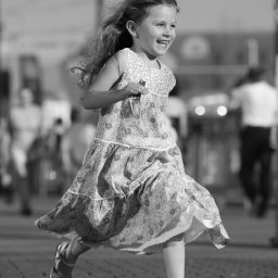 BRUNON FIDRYCH - Czarno-białe zdjęcie uśmiechniętej dziewczynki biegnącej po chodniku w kwiecistej sukience i białych sandałach, z rozpuszczonymi włosami powiewającymi na wietrze, na rozmytym tle miejskim.