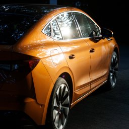 BRUNON FIDRYCH - Pomarańczowy samochód elektryczny Skoda Enyaq iV z aluminiowymi felgami, uchwycony w kontraście światła i cienia, z widocznym odbiciem słońca w szybie.