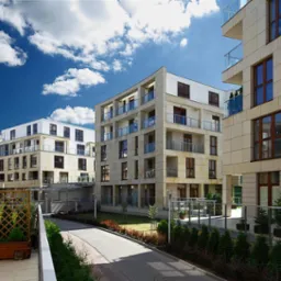 Nowoczesne budynki apartamentowe z elewacją z piaskowca, szklane balustrady balkon&oacute;w, zadbana zieleń, kostka brukowa i błękitne niebo z chmurami.