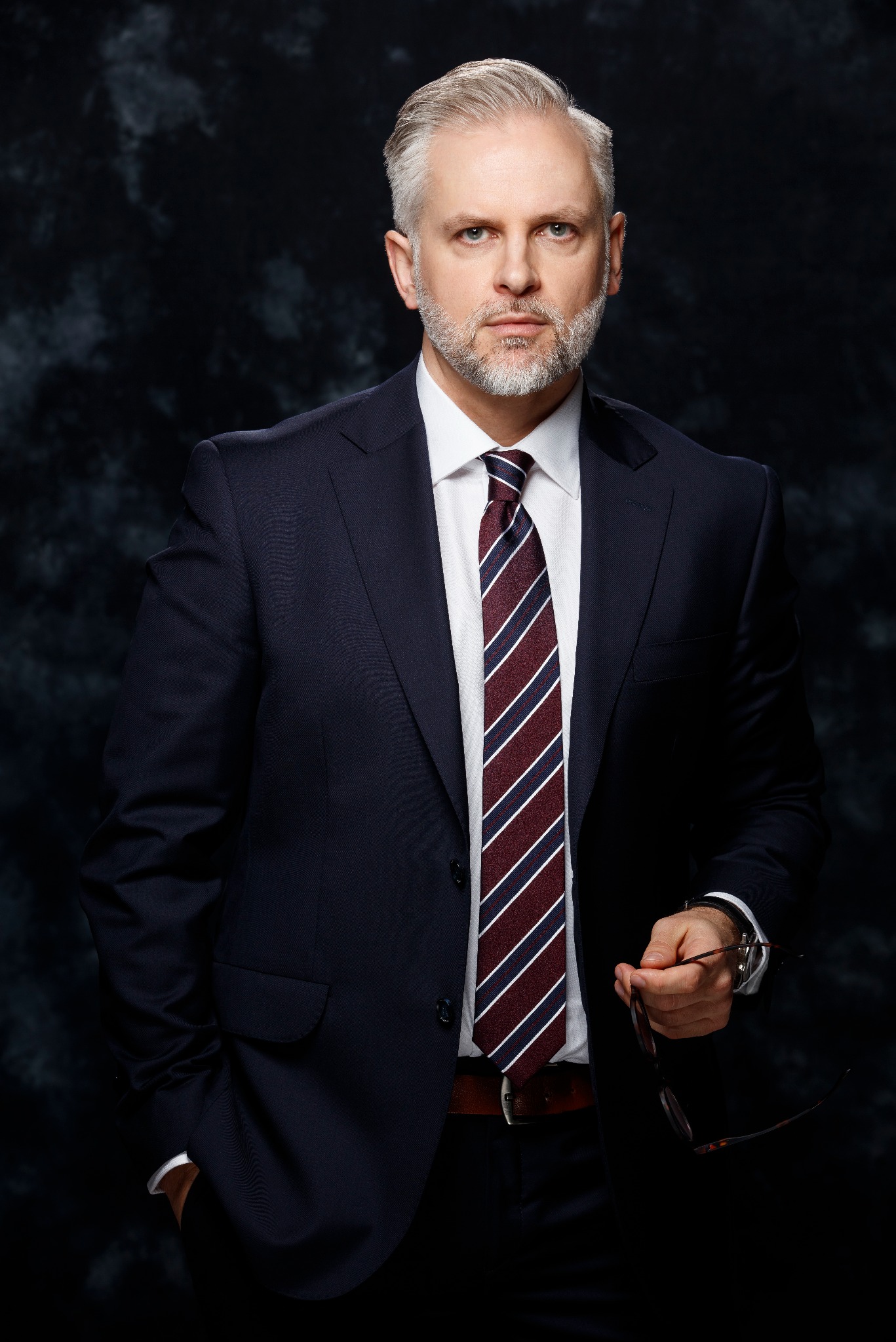 Portret biznesowy mężczyzny z siwymi włosami w granatowym garniturze i krawacie w paski, trzymającego okulary na tle ciemnego, teksturowanego tła.