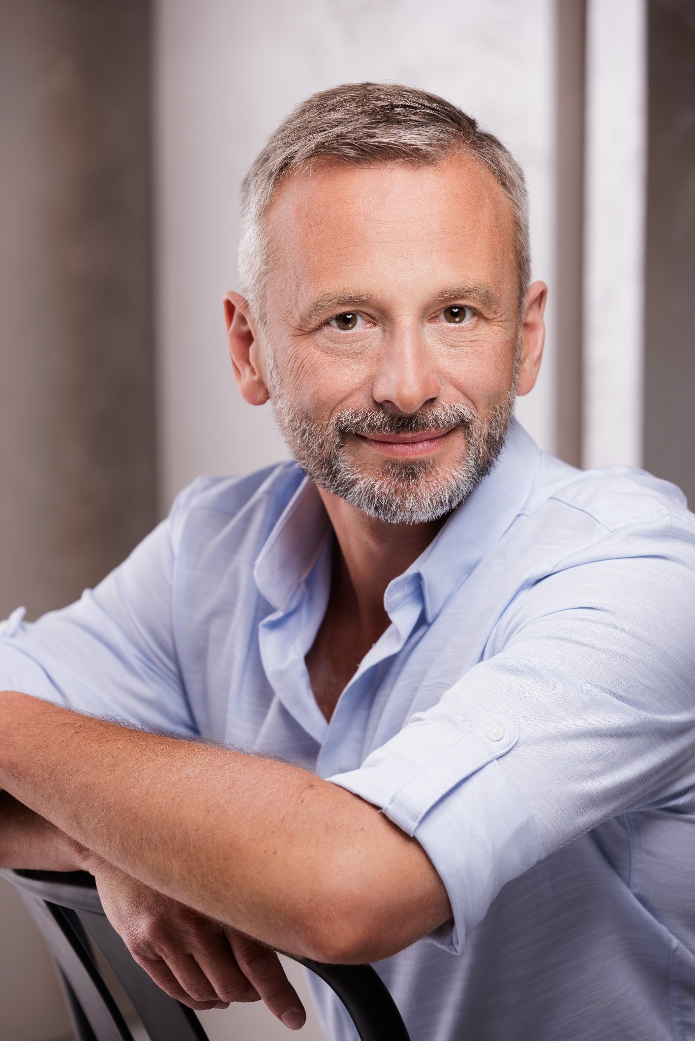 Portret uśmiechniętego, dojrzałego mężczyzny z siwiejącą brodą, w błękitnej koszuli, opierającego ręce na krześle, na neutralnym tle.