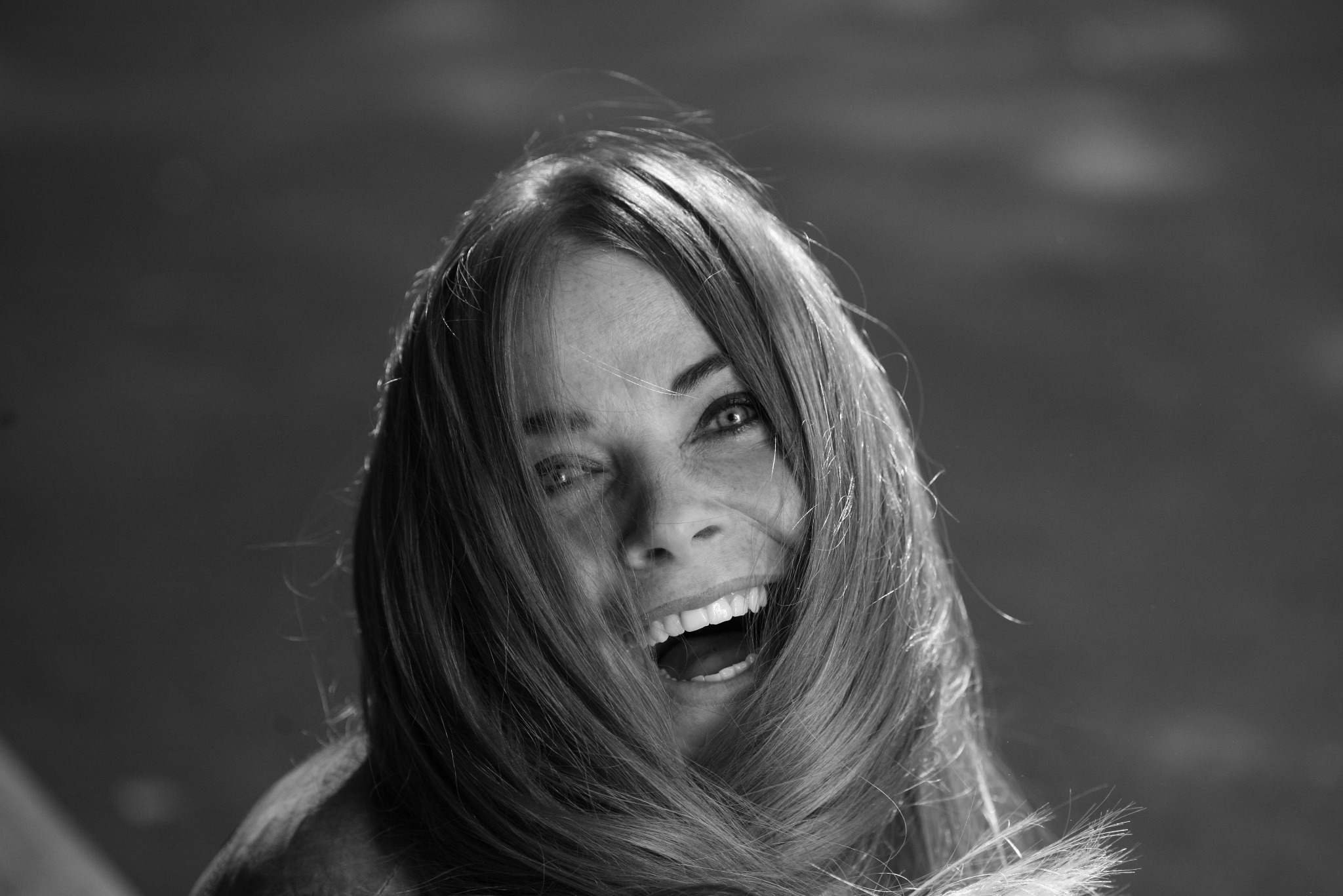 Portret roześmianej kobiety z rozwianymi włosami na wietrze, czarno-biały, z widocznymi detalami twarzy i włosów.