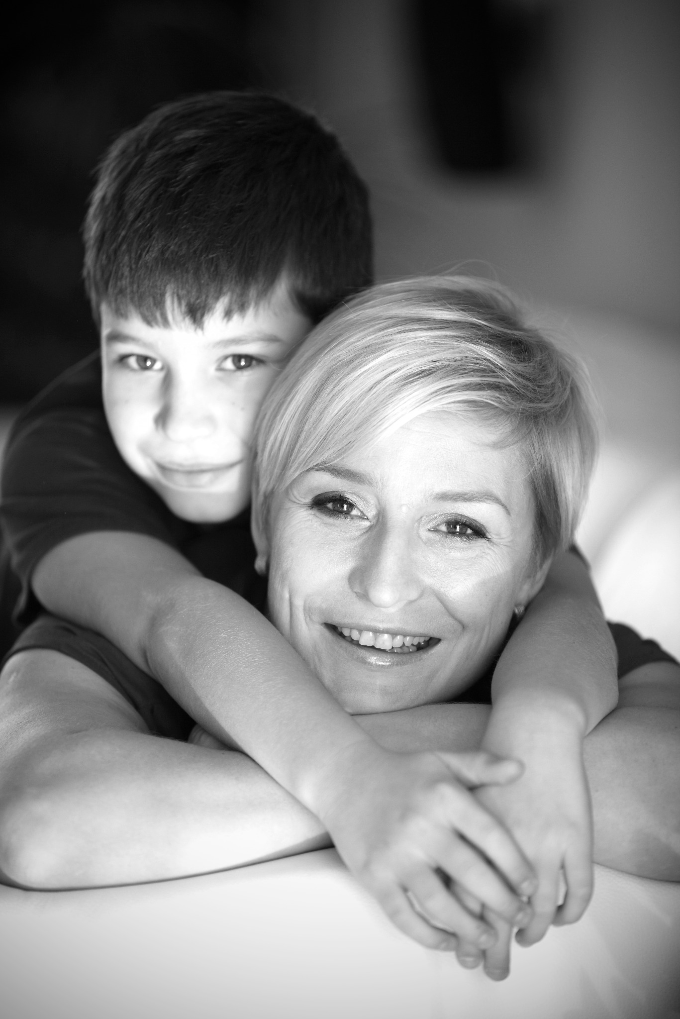 Czarno-białe zbliżenie uśmiechniętej kobiety z krótkimi blond włosami, obejmowanej od tyłu przez chłopca z ciemnymi włosami