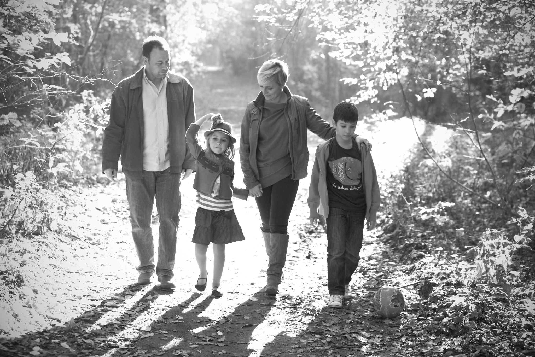 Czarno-białe zdjęcie rodziny spacerującej po lesie w słoneczny dzień; dziewczynka w kapeluszu podnosi rękę, a obok leży piłka.