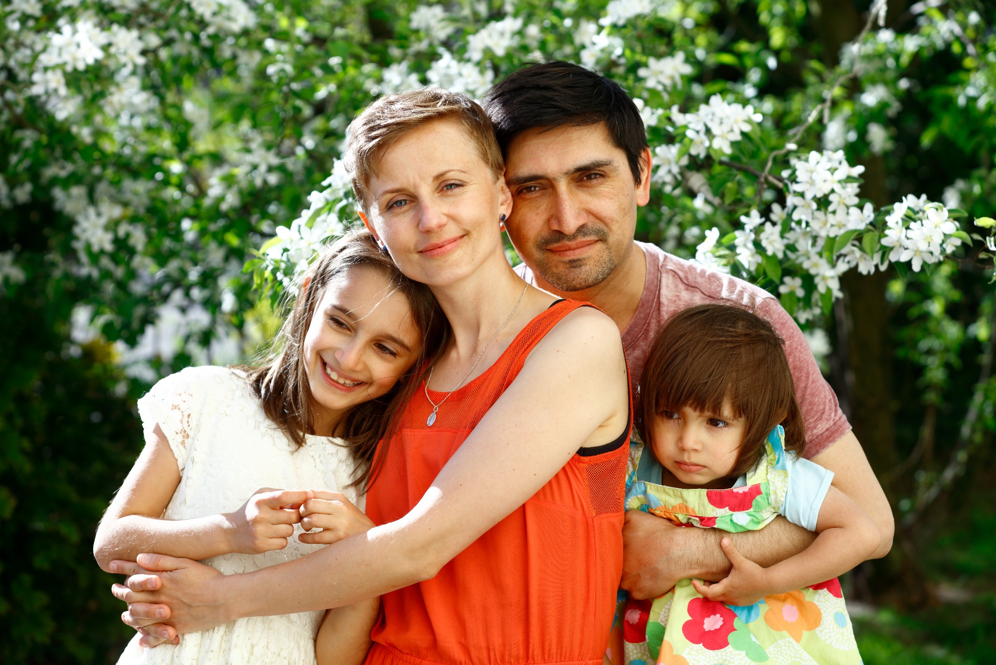 Uśmiechnięta rodzina: mama w pomarańczowej sukience, tata, córka w białej koronkowej sukience i młodsza córka w kolorowej sukience, pozują na tle kwitnącego drzewa.