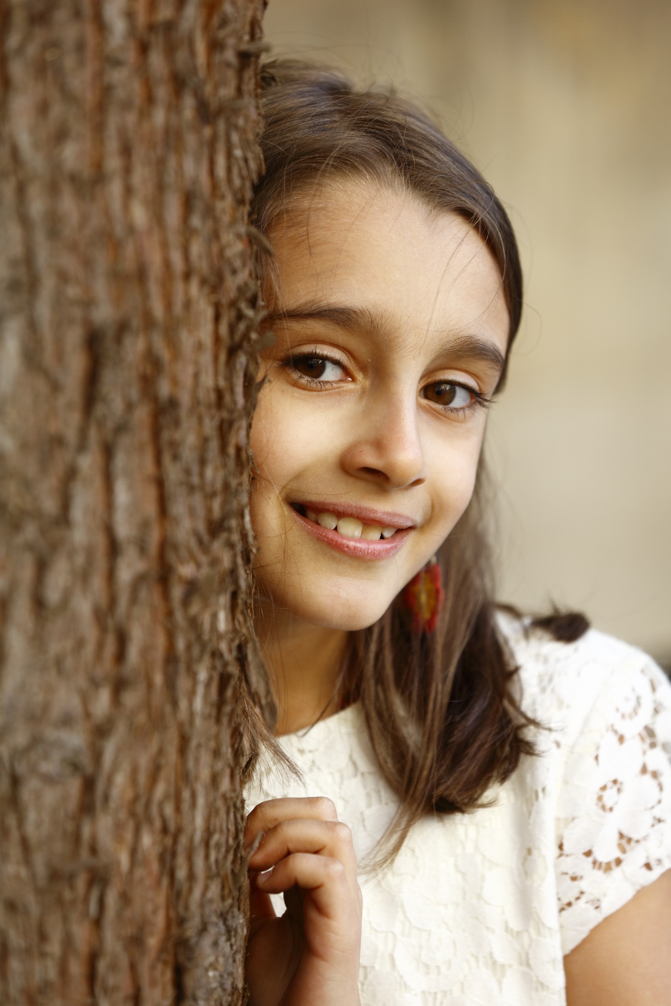 Portret uśmiechniętej dziewczynki w białej koronkowej bluzce, ukrywającej się częściowo za pniem drzewa z wyraźną fakturą kory, z widocznym kolczykiem w kształcie kwiatu.