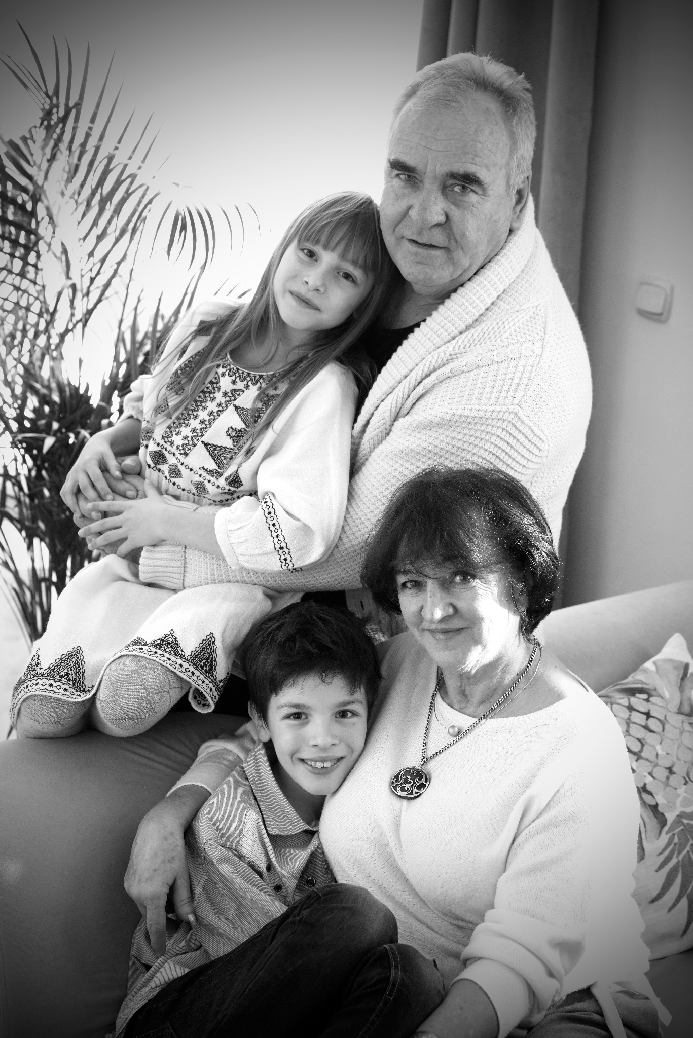 Czarno-białe portretowe ujęcie czterech osób z rodziny: dziadek w swetrze, babcia z naszyjnikiem oraz dwoje wnucząt w tradycyjnych strojach z haftami, siedzących na kanapie na tle rośliny doniczkowej.