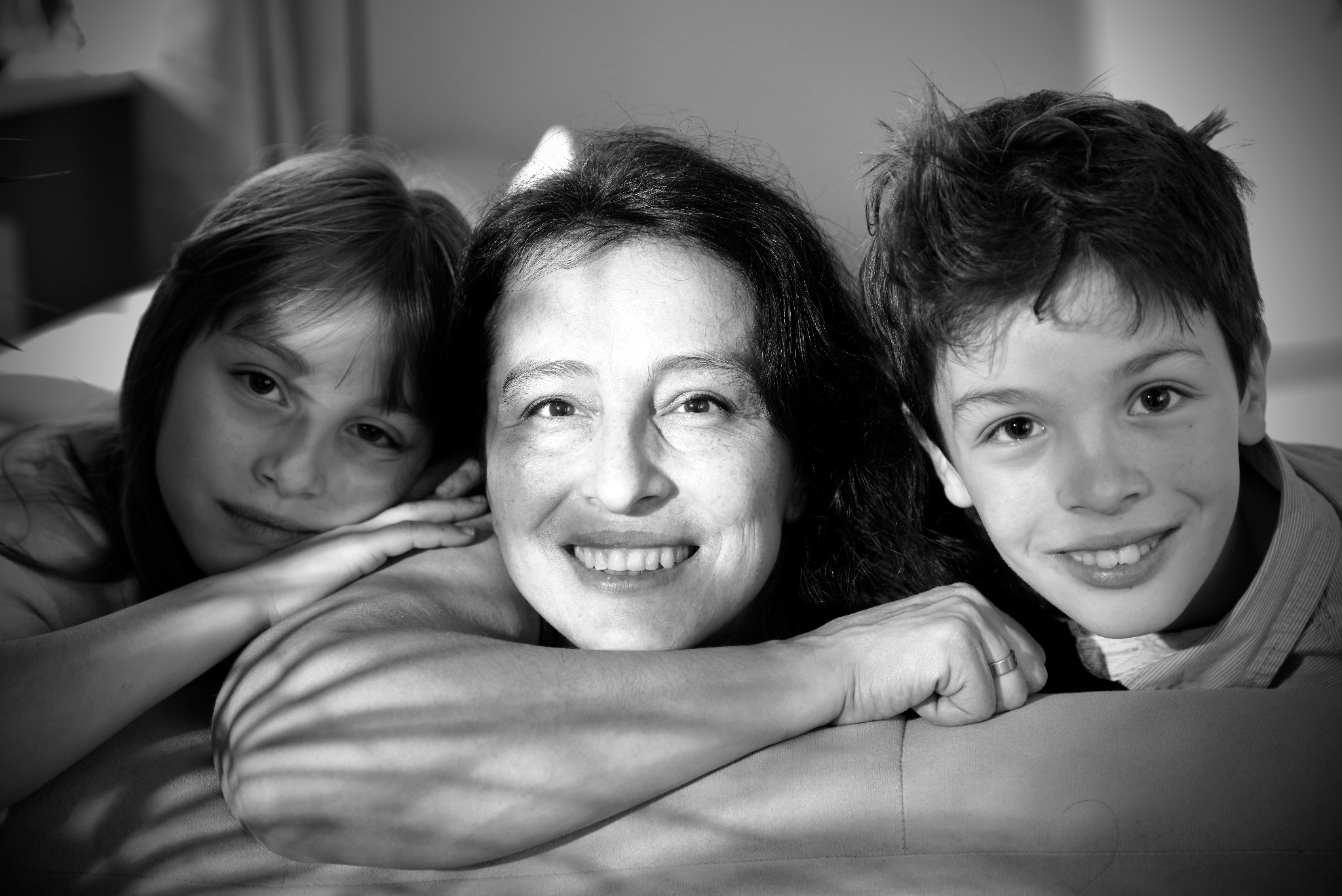 Czarno-białe portretowe ujęcie matki z dwójką dzieci, uśmiechniętych i opartych na rękach, z delikatnymi cieniami na skórze.