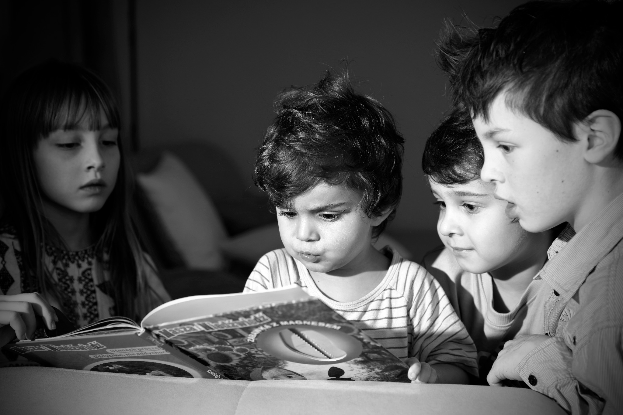Czarno-białe zdjęcie czwórki dzieci skupionych na czytaniu książki z magnetyczną zabawką, dziewczynka z długimi włosami po lewej, chłopiec w paski na środku, dwaj chłopcy w koszulach po prawej.