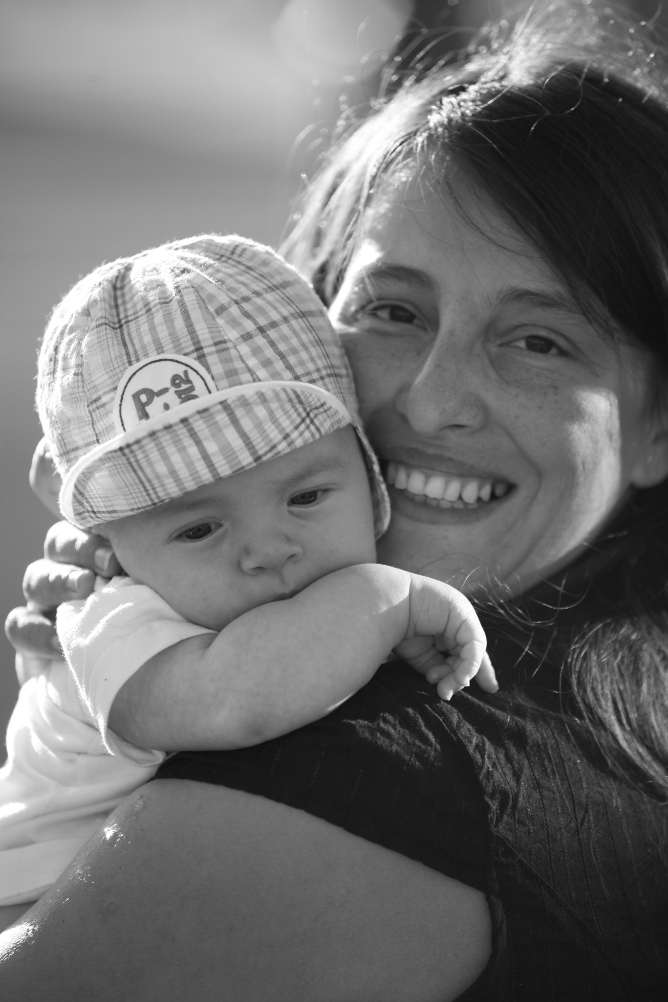 Czarno-białe zdjęcie uśmiechniętej kobiety trzymającej na rękach niemowlę w czapce w kratę w Warszawie.