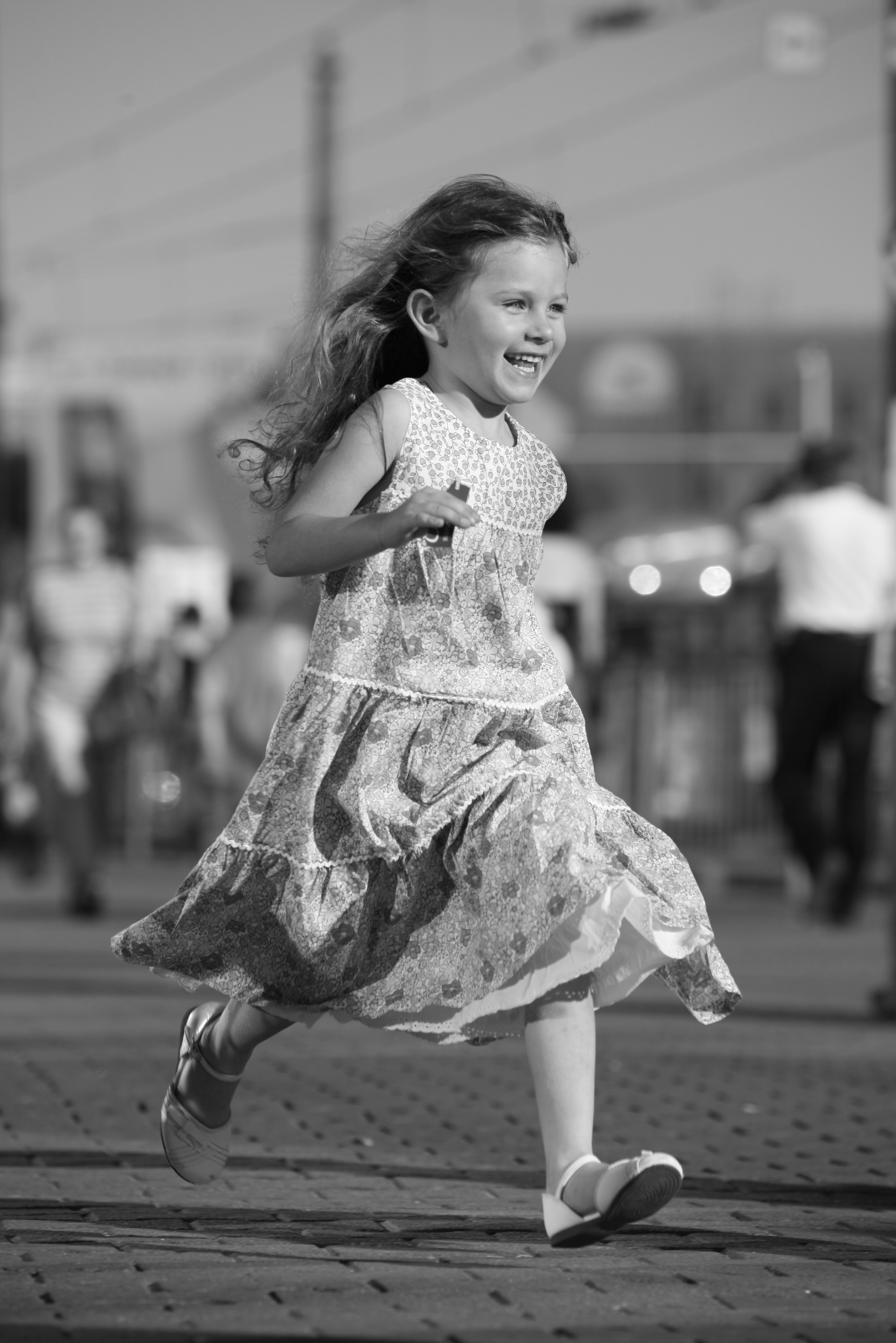 Czarno-białe zdjęcie uśmiechniętej dziewczynki biegnącej po chodniku w kwiecistej sukience i białych sandałach, z rozpuszczonymi włosami powiewającymi na wietrze, na rozmytym tle miejskim.