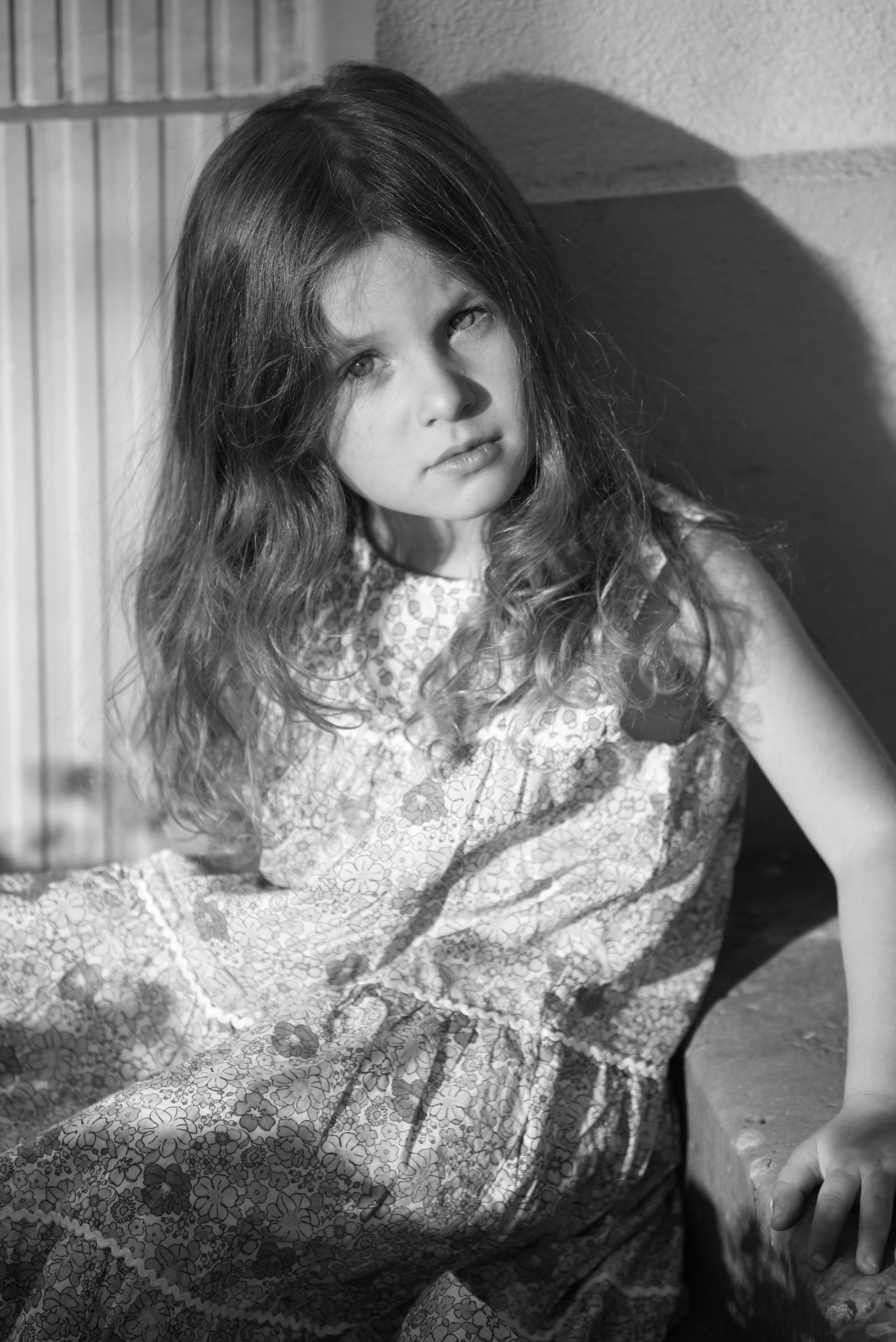 Portret czarno-biały dziewczynki w kwiecistej sukience, oparta o betonową ścianę, oświetlona bocznym światłem.