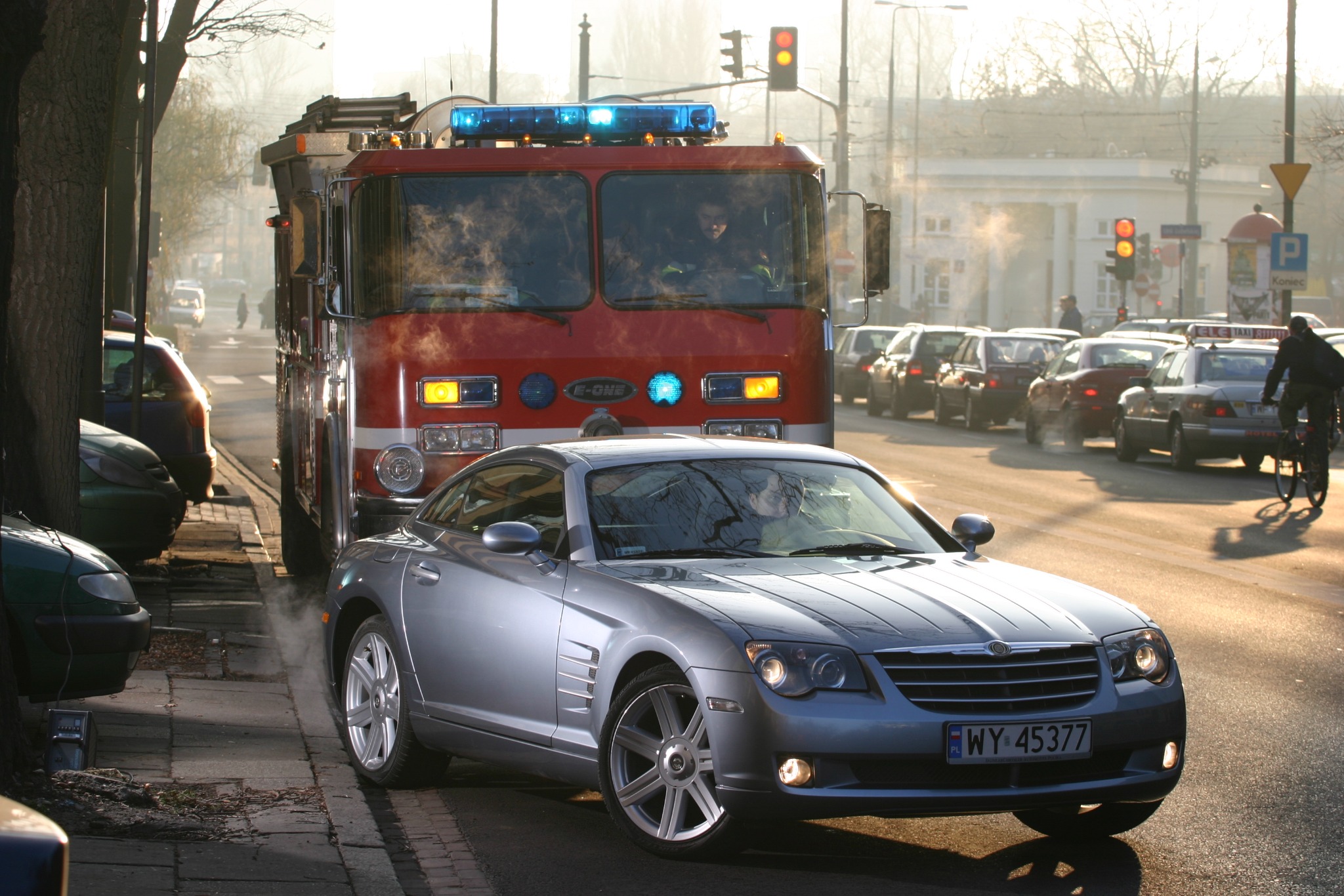 Srebrny Chrysler Crossfire zaparkowany na chodniku obok wozu strażackiego na tle ulicy miejskiej z sygnalizacją świetlną.