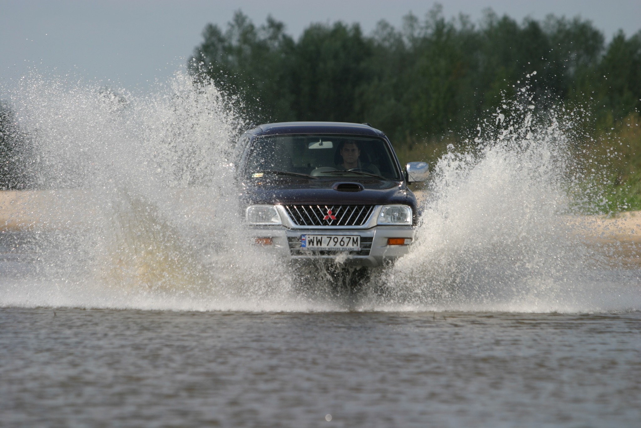 Ciemny samochód terenowy Mitsubishi przedziera się przez wodę, tworząc fontannę rozprysków, widok z przodu, drzewa w tle.
