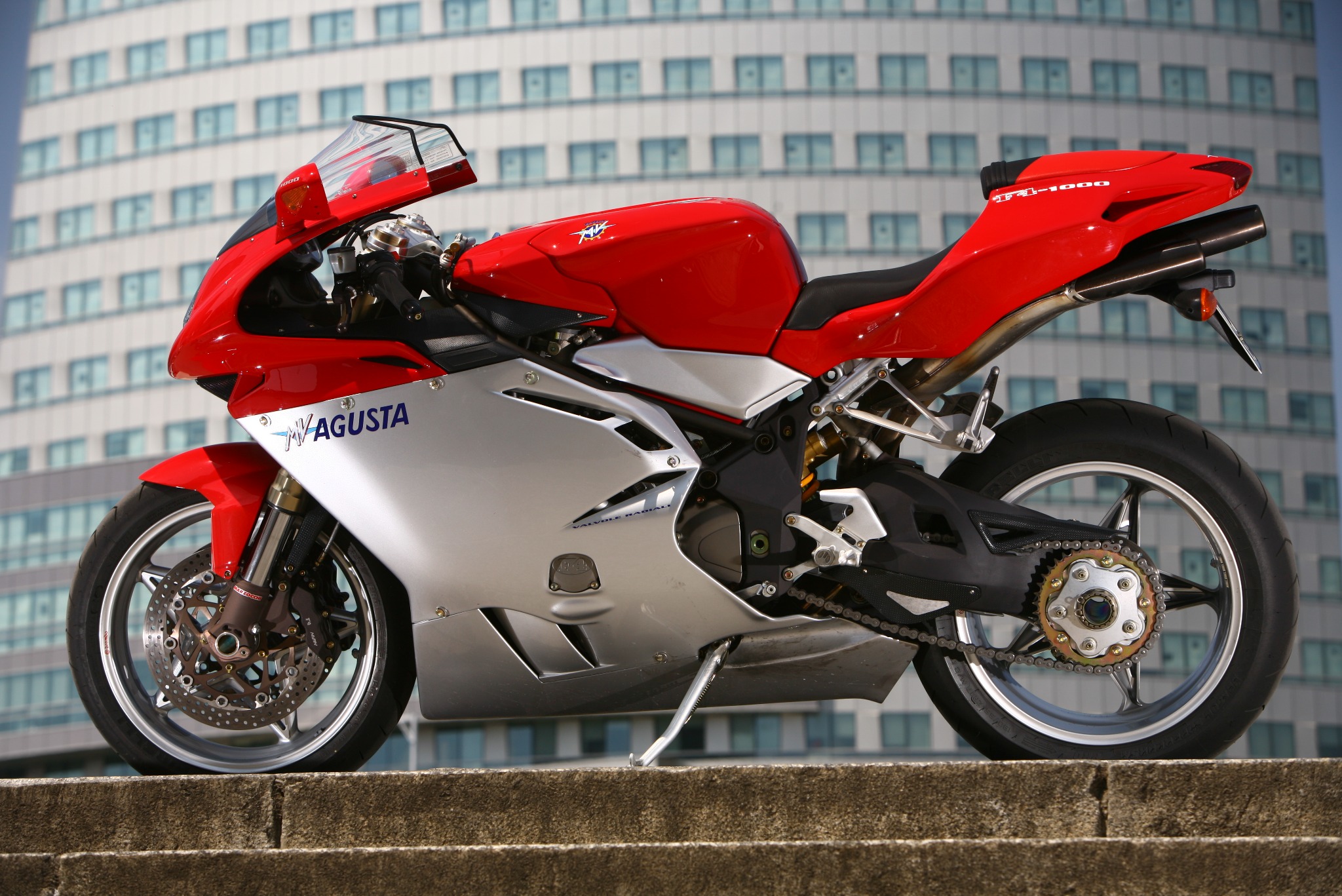 Czerwono-srebrny motocykl MV Agusta F4 1000 zaparkowany na betonowych schodach z nowoczesnym budynkiem w tle.