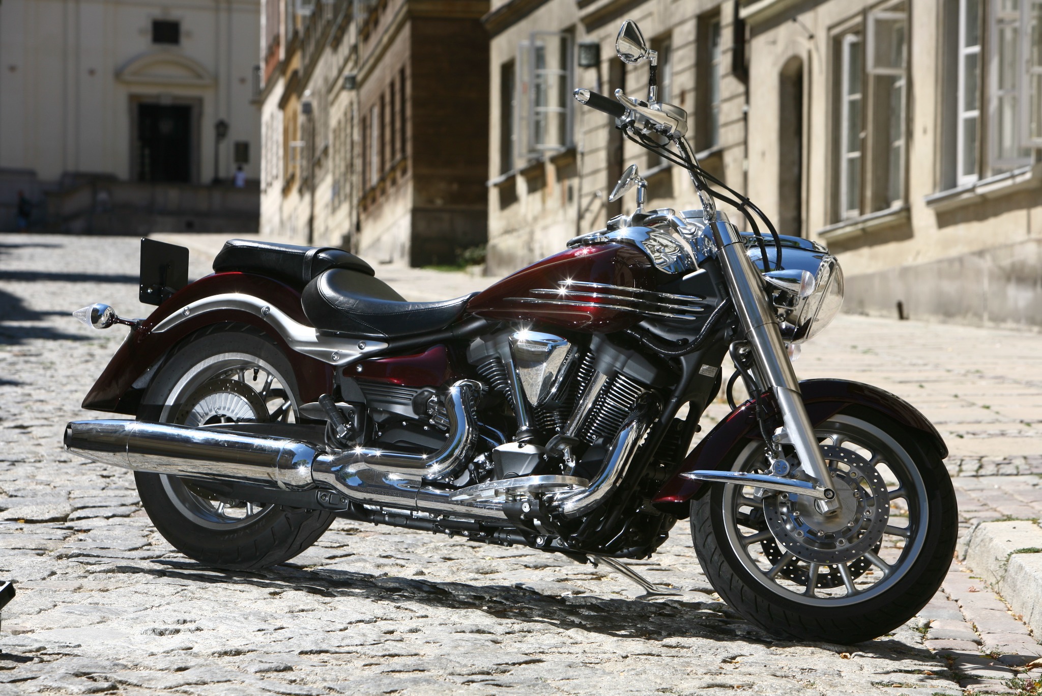 Bordowy motocykl cruiser z chromowanymi elementami zaparkowany na brukowanej ulicy w słoneczny dzień, budynki w tle.