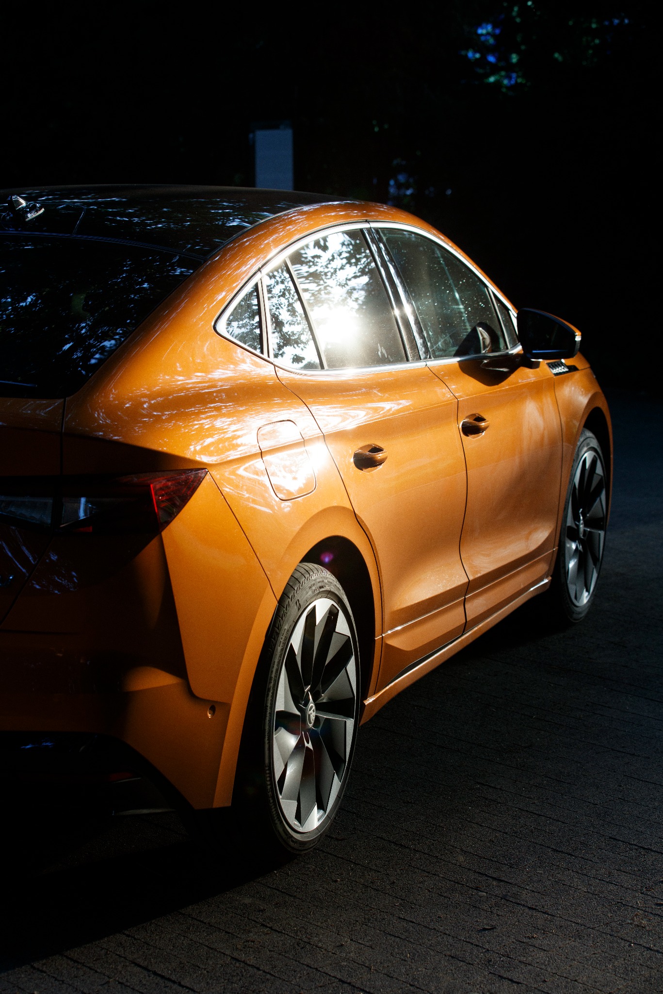 Pomarańczowy samochód elektryczny Skoda Enyaq iV z aluminiowymi felgami, uchwycony w kontraście światła i cienia, z widocznym odbiciem słońca w szybie.