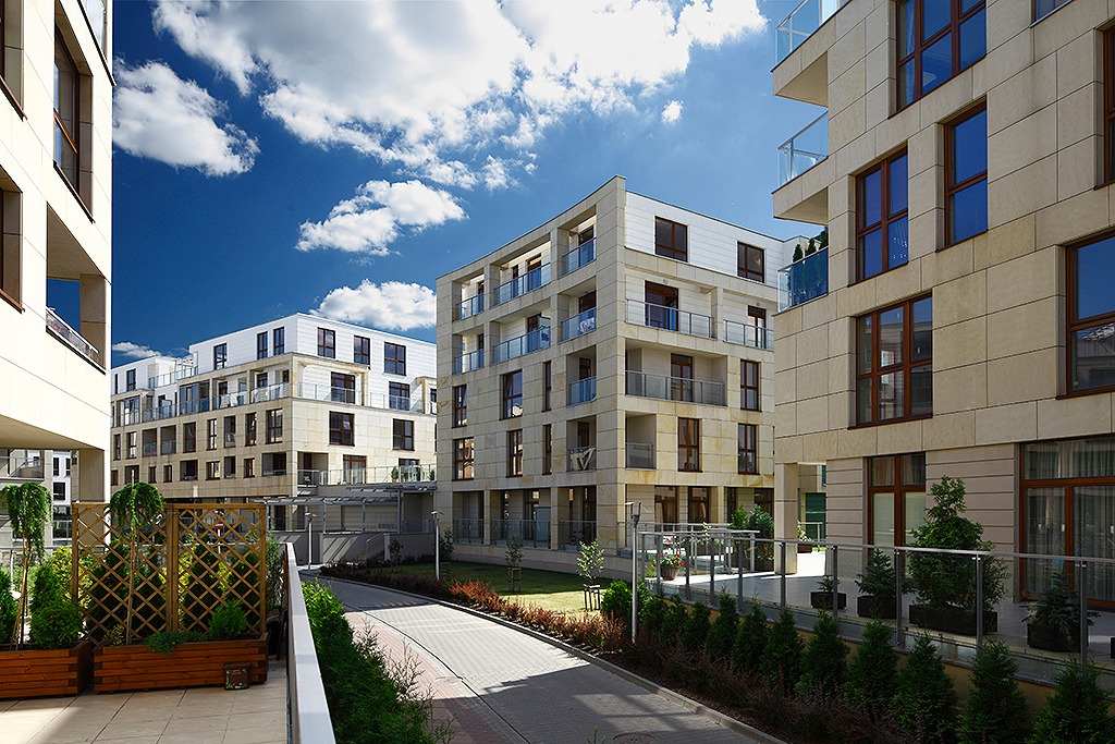 Nowoczesne budynki apartamentowe z elewacją z piaskowca, szklane balustrady balkonów, zadbana zieleń, kostka brukowa i błękitne niebo z chmurami.