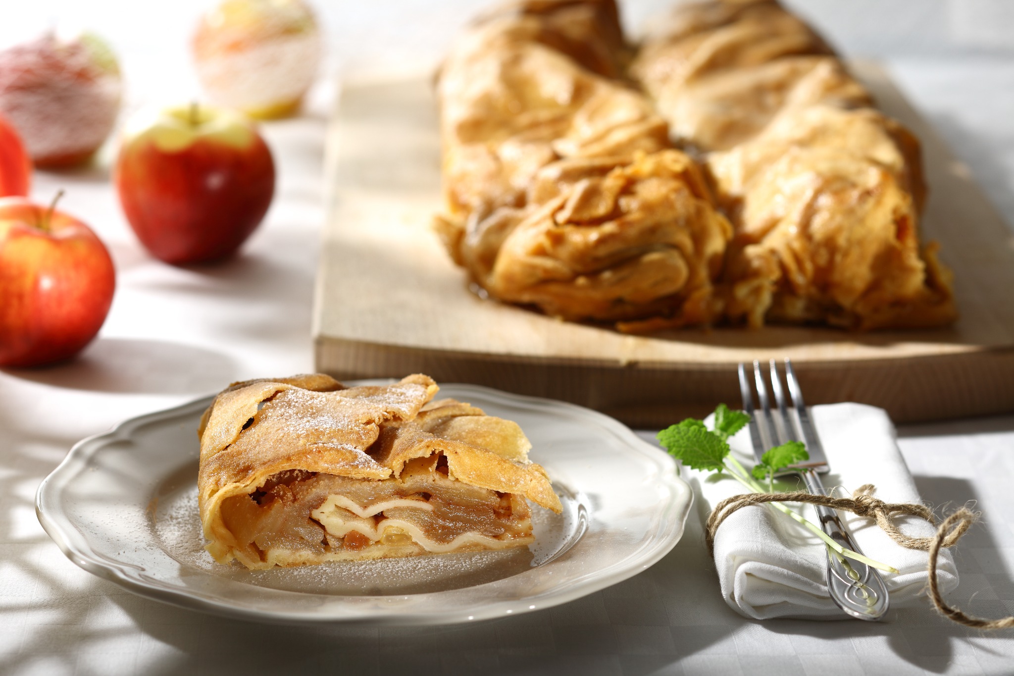 Apetyczny kawałek szarlotki na białym talerzu, posypany cukrem pudrem, z jabłkami w tle i widelcem owiniętym sznurkiem jutowym na serwetce.