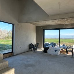 Adrian Ślipski - Surowe wnętrze domu w trakcie tynkowania maszynowego; widoczne gładkie, szare ściany, betonowe schody, duże okna z widokiem na zielone pole i drzewa, worki z materiałami budowlanymi i plastikowe...