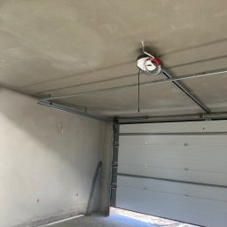Adrian Ślipski - Surowy garaż z świeżo nałożonym tynkiem na ścianach i suficie, widoczny mechanizm otwierania bramy garażowej, brama częściowo otwarta.