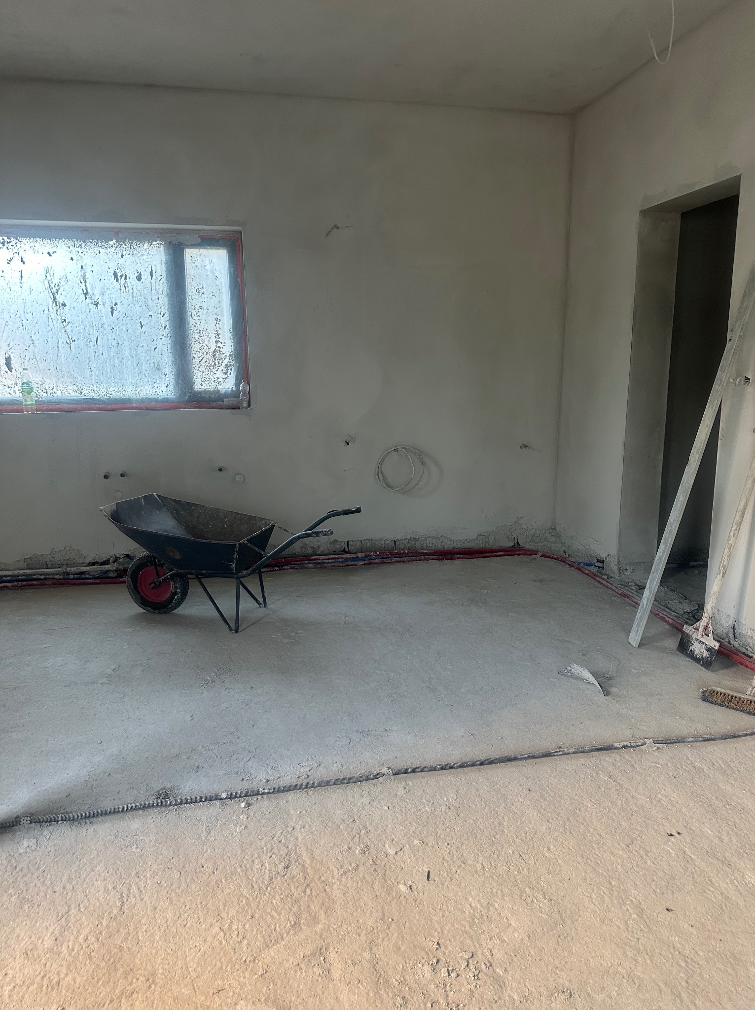 Wnętrze w trakcie tynkowania maszynowego: widoczna taczka z resztkami zaprawy, ściany z nałożonym tynkiem, instalacja wodna przy podłodze i brudne okno.