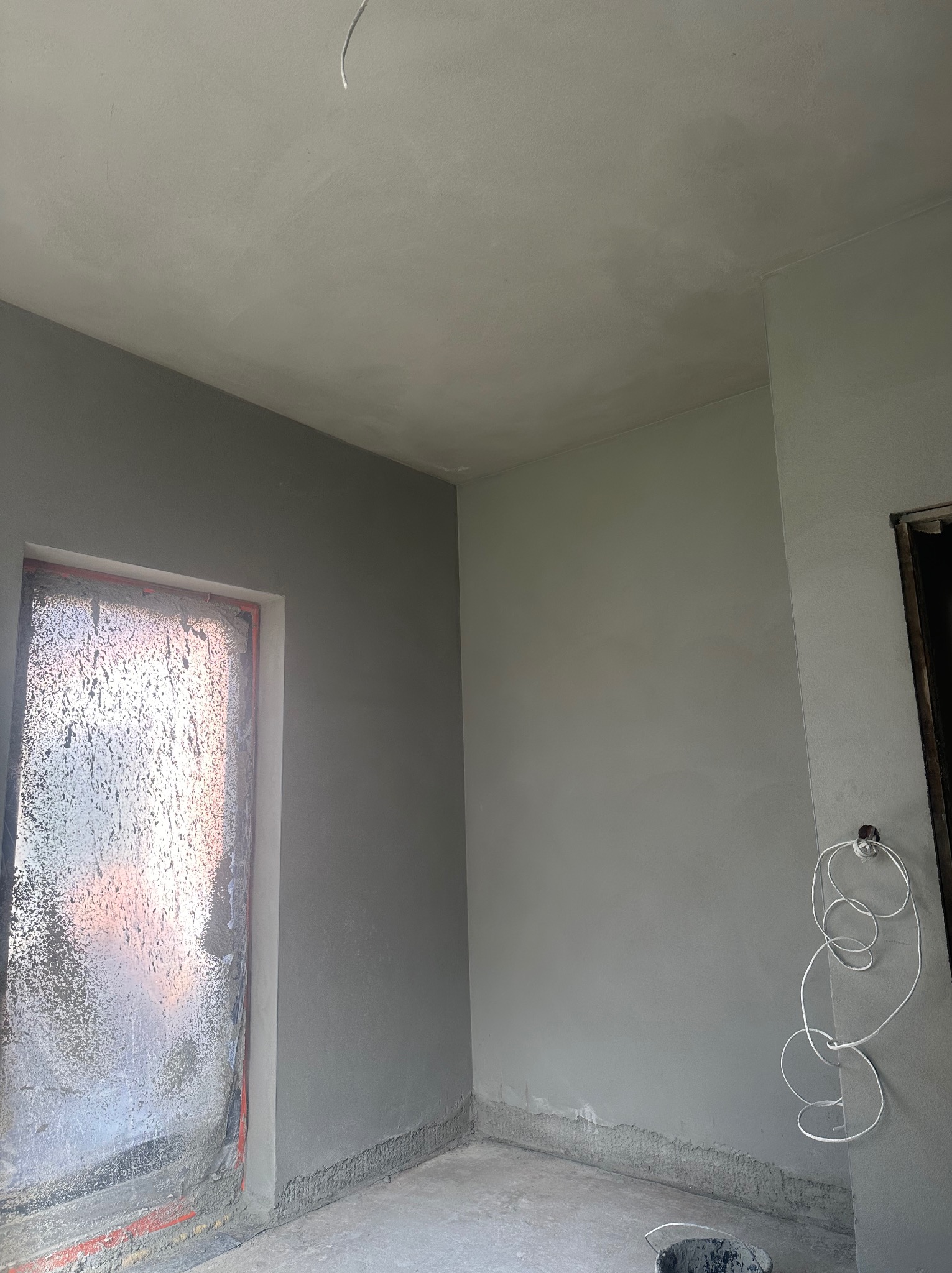 Świeżo położony szary tynk na ścianach i suficie w pomieszczeniu w trakcie remontu, okno zabezpieczone folią, widoczna instalacja elektryczna.