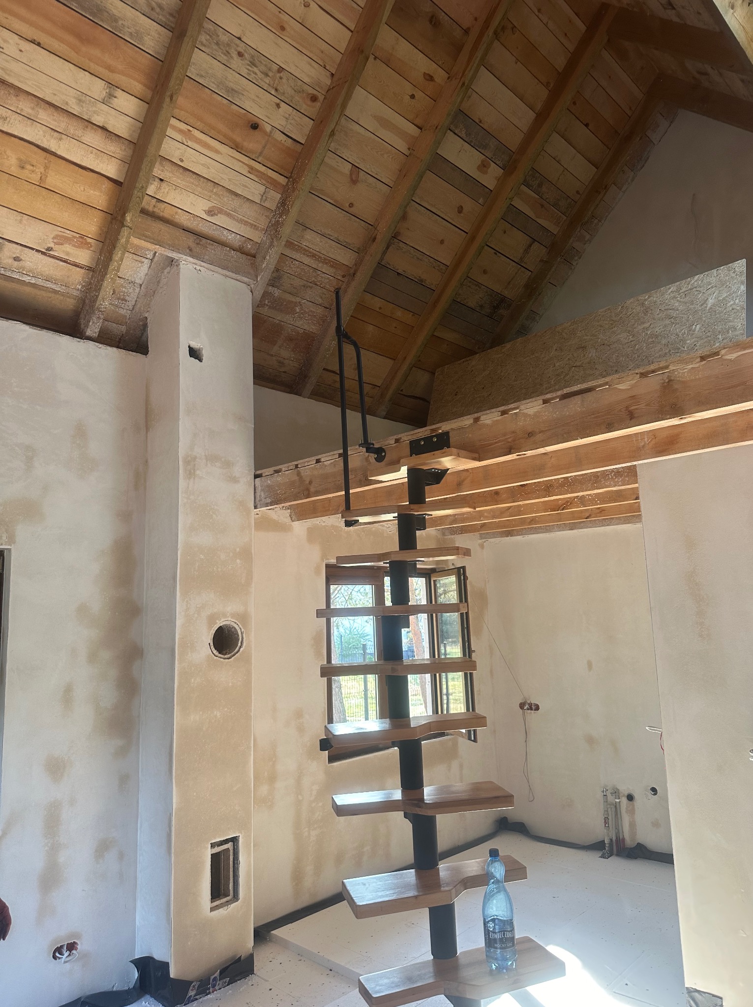 Wnętrze w trakcie wykończenia: surowy drewniany strop, ściany po nałożeniu pierwszej warstwy gładzi gipsowej, spiralne schody z drewnianymi stopniami i czarną metalową konstrukcją, butelka wody...