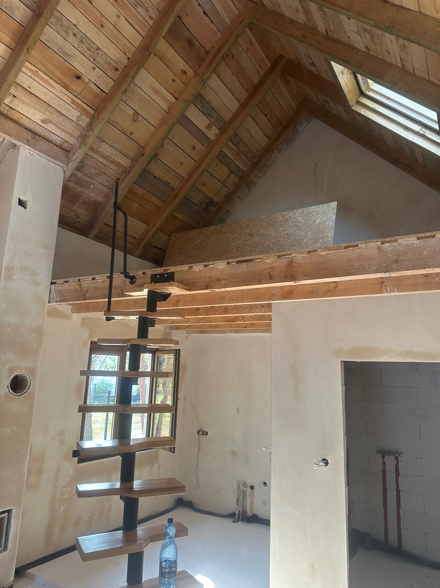 Wnętrze poddasza w trakcie wykańczania: surowe drewniane deski na suficie, spiralne schody z drewnianymi stopniami, ściany po wstępnym nałożeniu tynku, widoczne elementy instalacji elektrycznej...