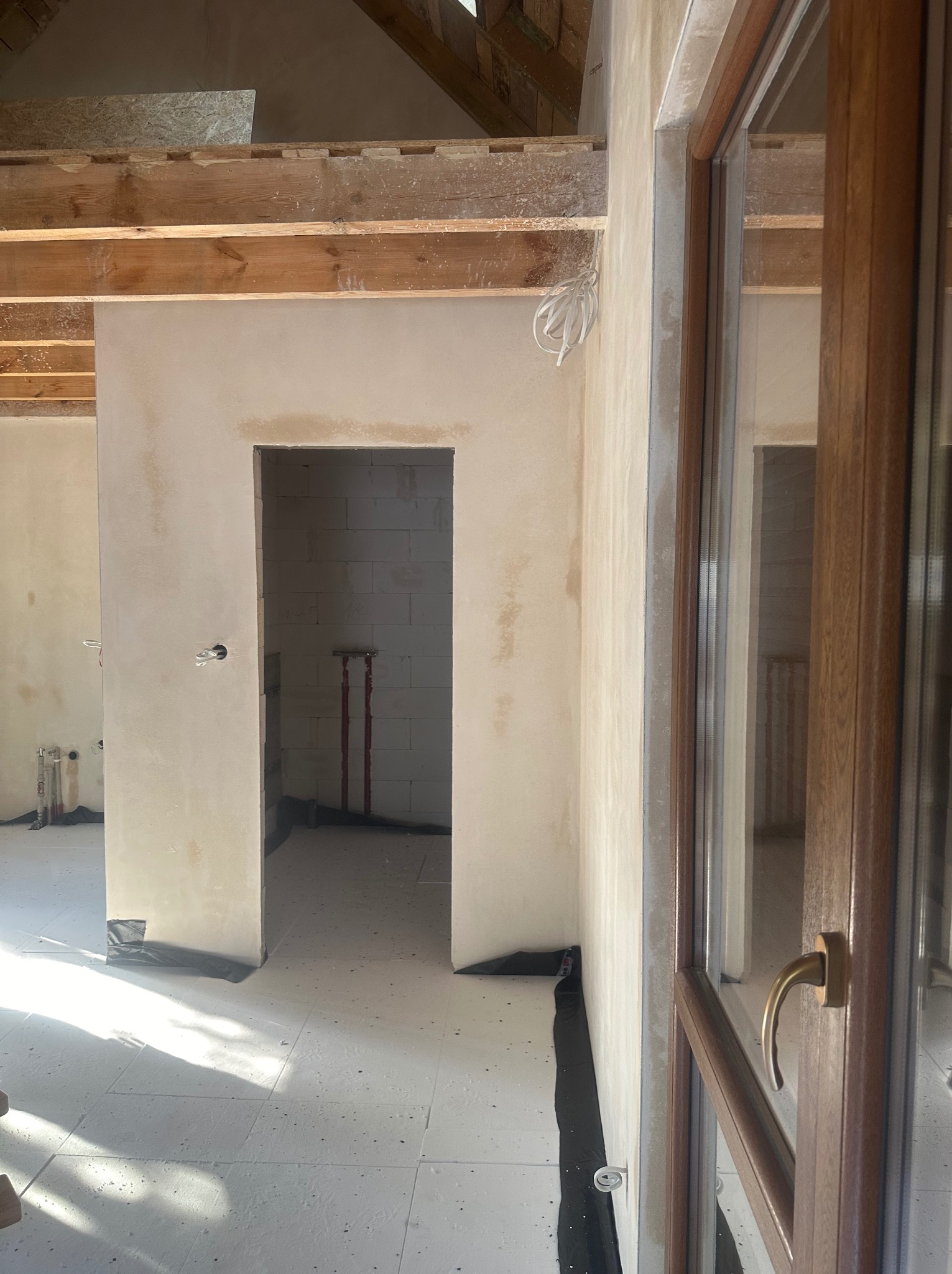 Surowe wnętrze w trakcie prac wykończeniowych, z widocznymi elementami konstrukcji drewnianej stropu, ścianami pokrytymi świeżym tynkiem i zabezpieczoną podłogą.