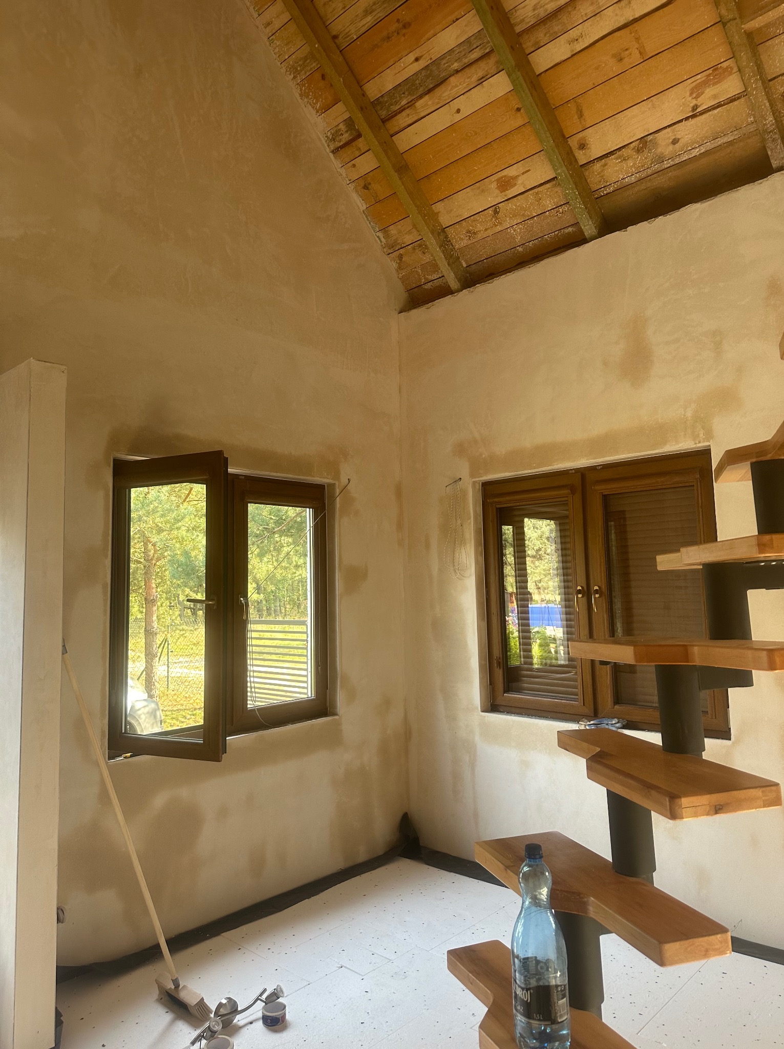 Wnętrze w trakcie prac wykończeniowych: ściany pokryte świeżym tynkiem, drewniane okna, surowe drewniane belki stropowe, tymczasowe schody drewniane, butelka wody na schodach, widoczne narzędzia...