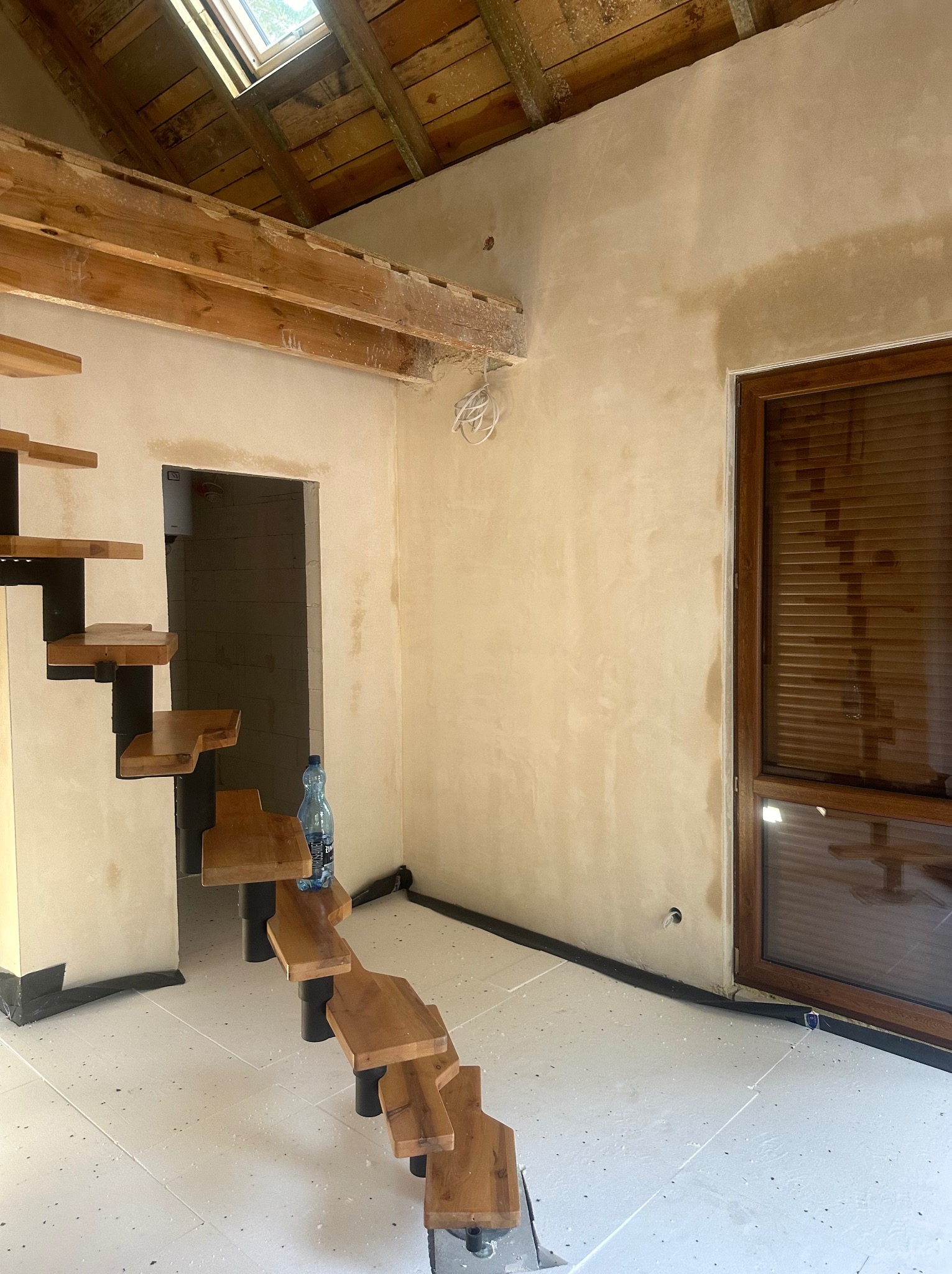 Wnętrze w trakcie wykańczania: ściana po nałożeniu gładzi, drewniane schody spiralne z czarnymi elementami, widoczne fragmenty drewnianej konstrukcji dachu i okno dachowe, butelka wody mineralnej...