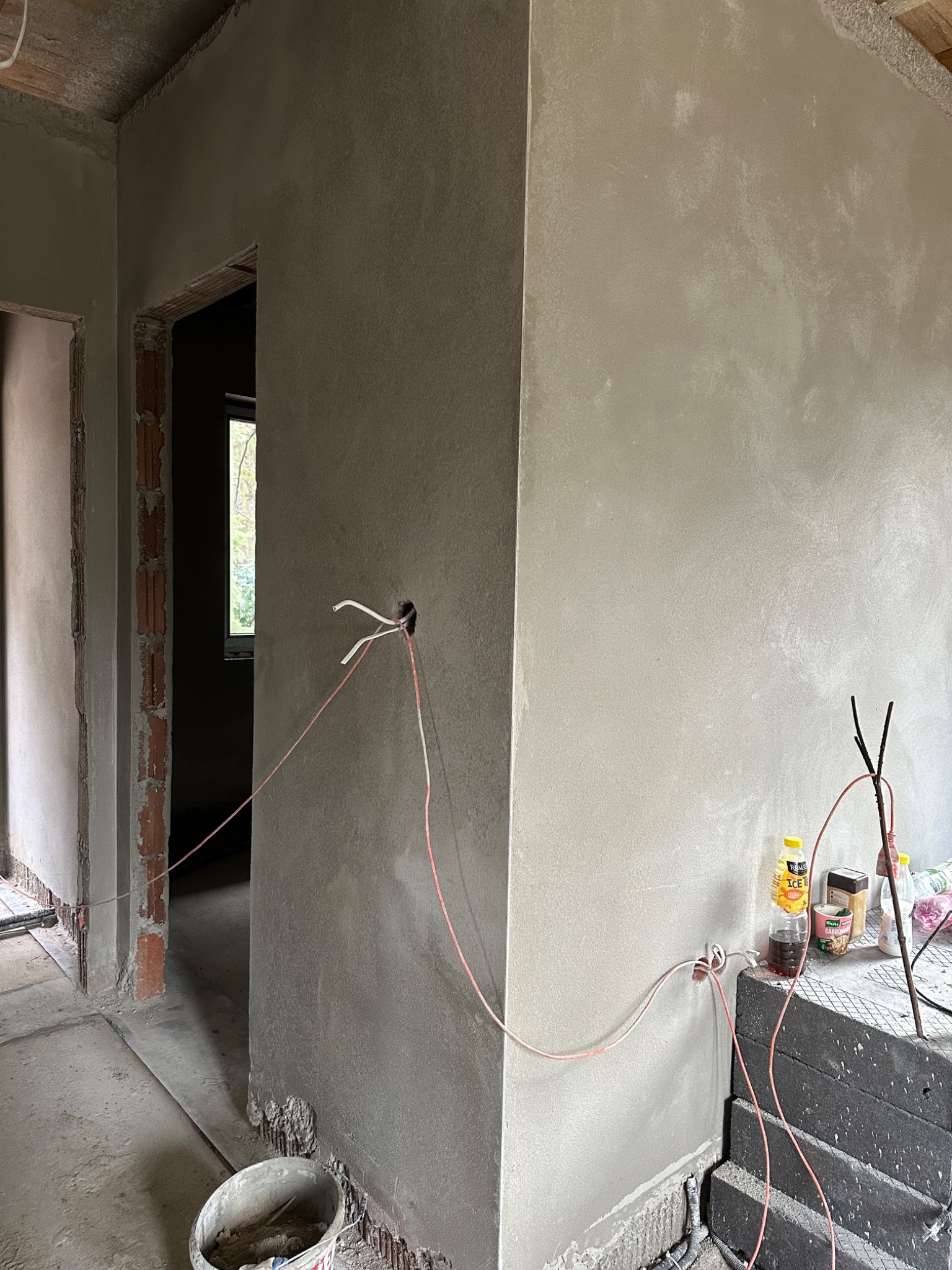 Wnętrze w trakcie prac wykończeniowych: świeżo otynkowane ściany z widocznymi przewodami elektrycznymi, fragmenty cegieł w otworach drzwiowych oraz wiadro z zaprawą na podłodze.