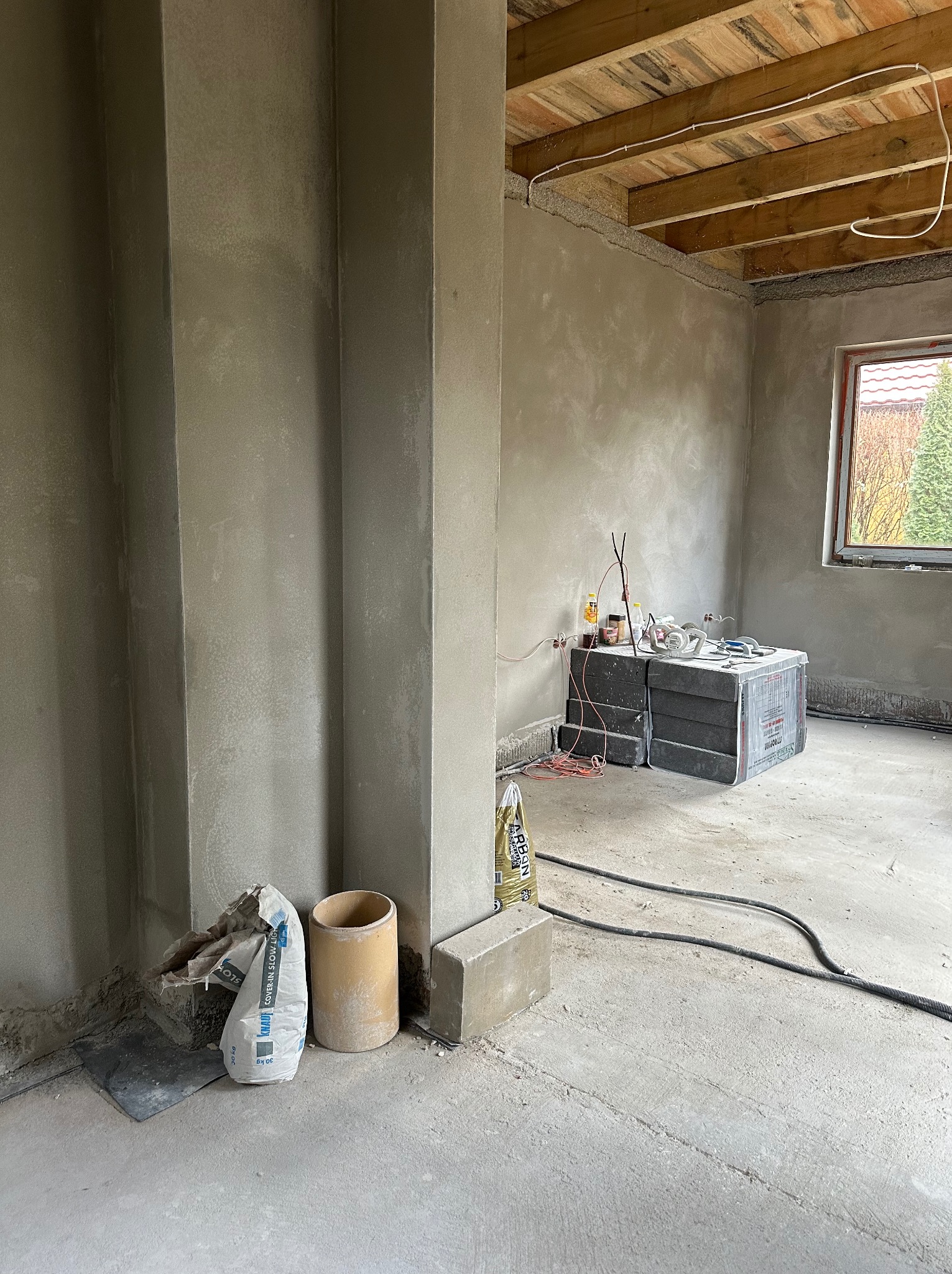 Wnętrze w trakcie prac wykończeniowych, ściany pokryte świeżym tynkiem, widoczne worki z zaprawą, narzędzia i materiały budowlane, surowy drewniany strop.