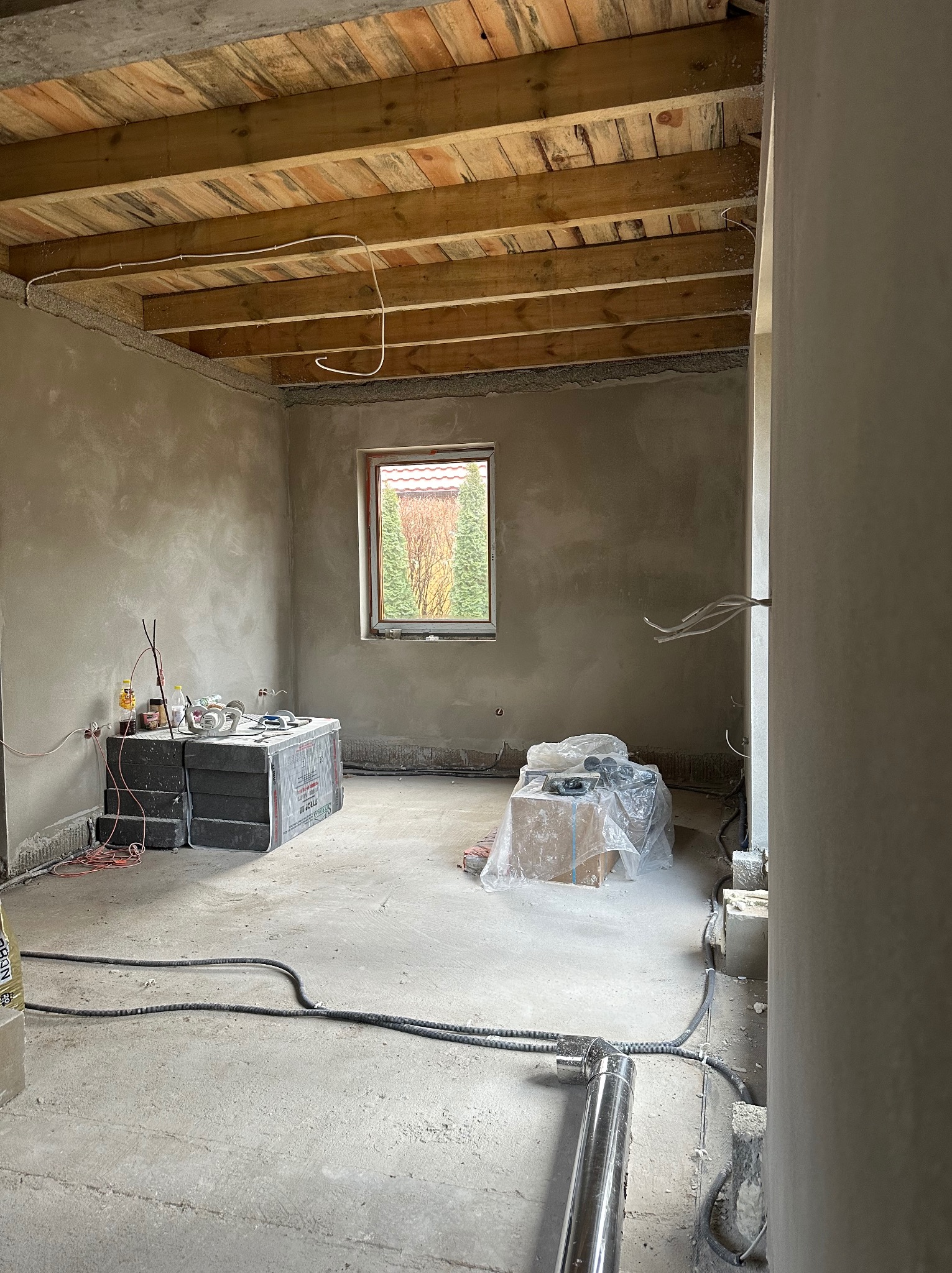Wnętrze surowego pomieszczenia po tynkowaniu maszynowym, z widoczną drewnianą konstrukcją stropu, przewodami elektrycznymi, oknem z widokiem na zieleń i materiałami budowlanymi na podłodze.
