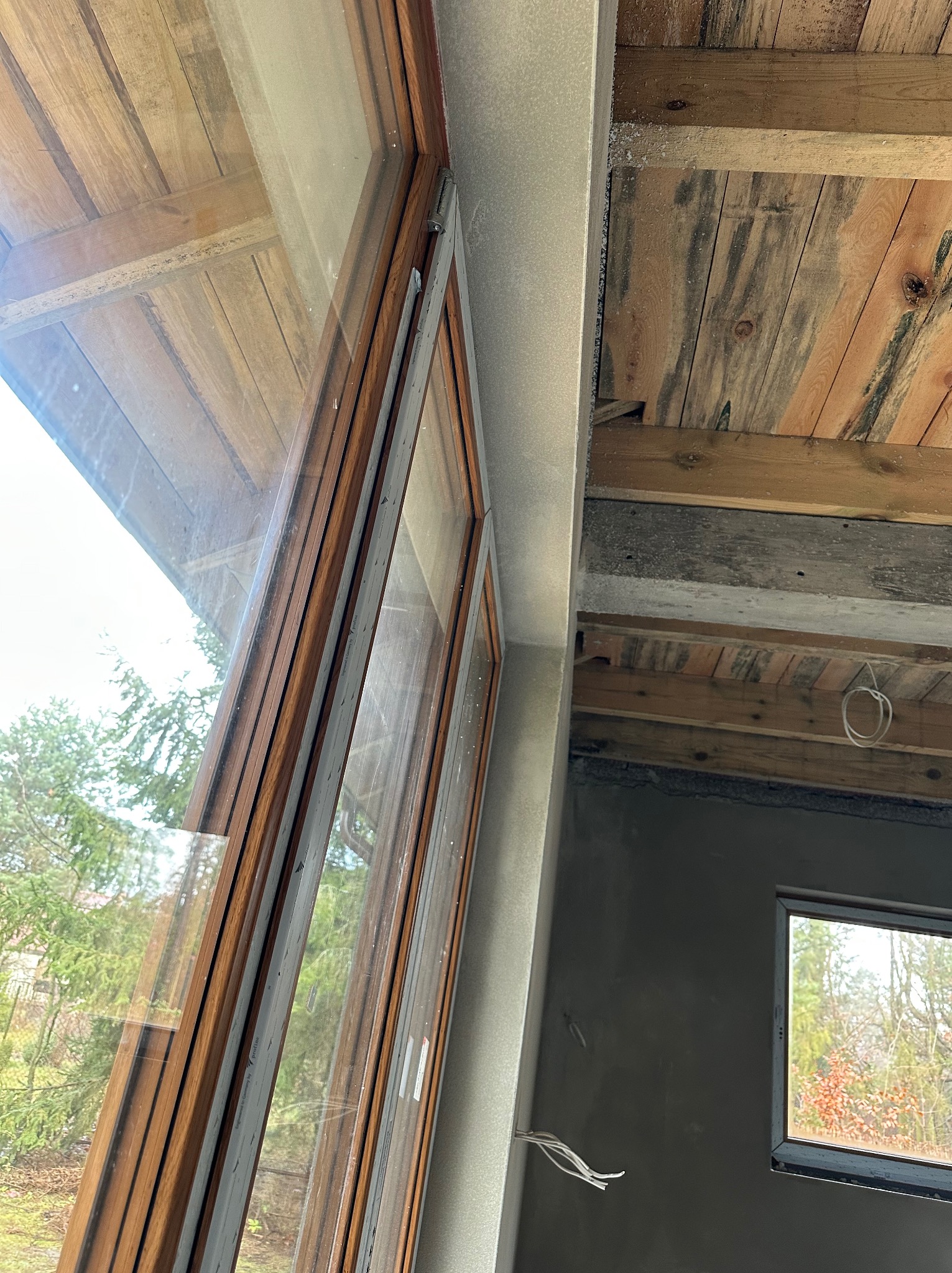 Wnętrze budynku w trakcie prac wykończeniowych: widoczne okno z drewnianą ramą, ściana pokryta świeżym, szarym tynkiem oraz surowy, drewniany strop z widocznymi belkami.