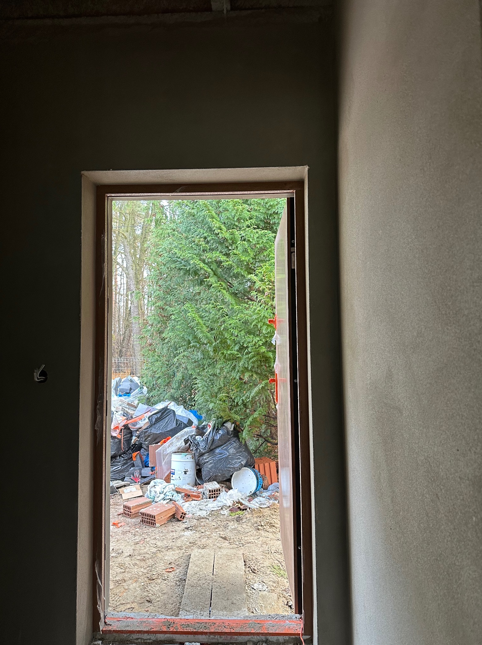 Surowa ściana z widocznym otworem drzwiowym, przez który widać gruz, cegły i worki na śmieci na zewnątrz.