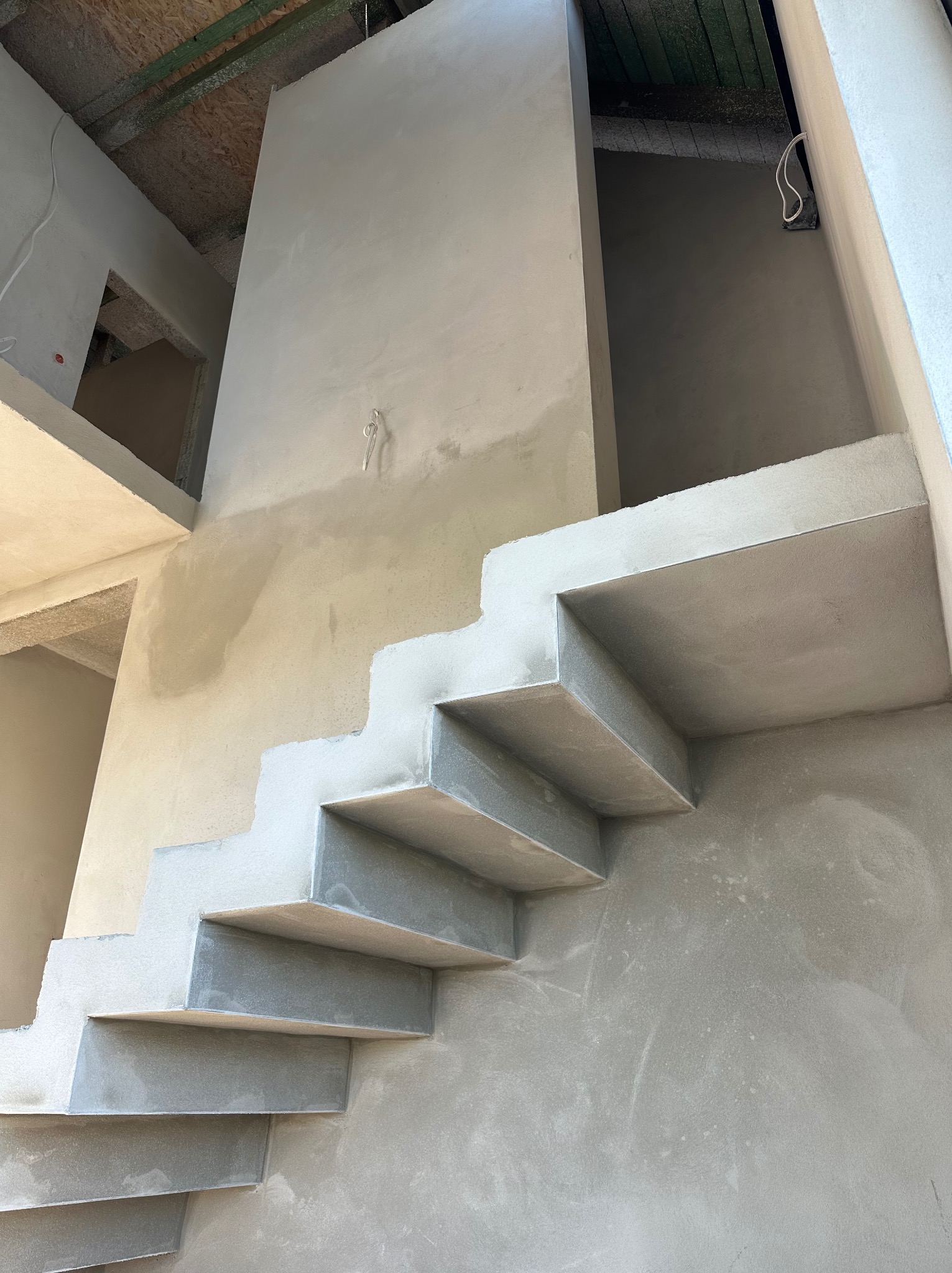 Surowe schody żelbetowe z widoczną strukturą świeżego tynku na ścianach w surowym stanie budynku, z widoczną instalacją elektryczną na górze.