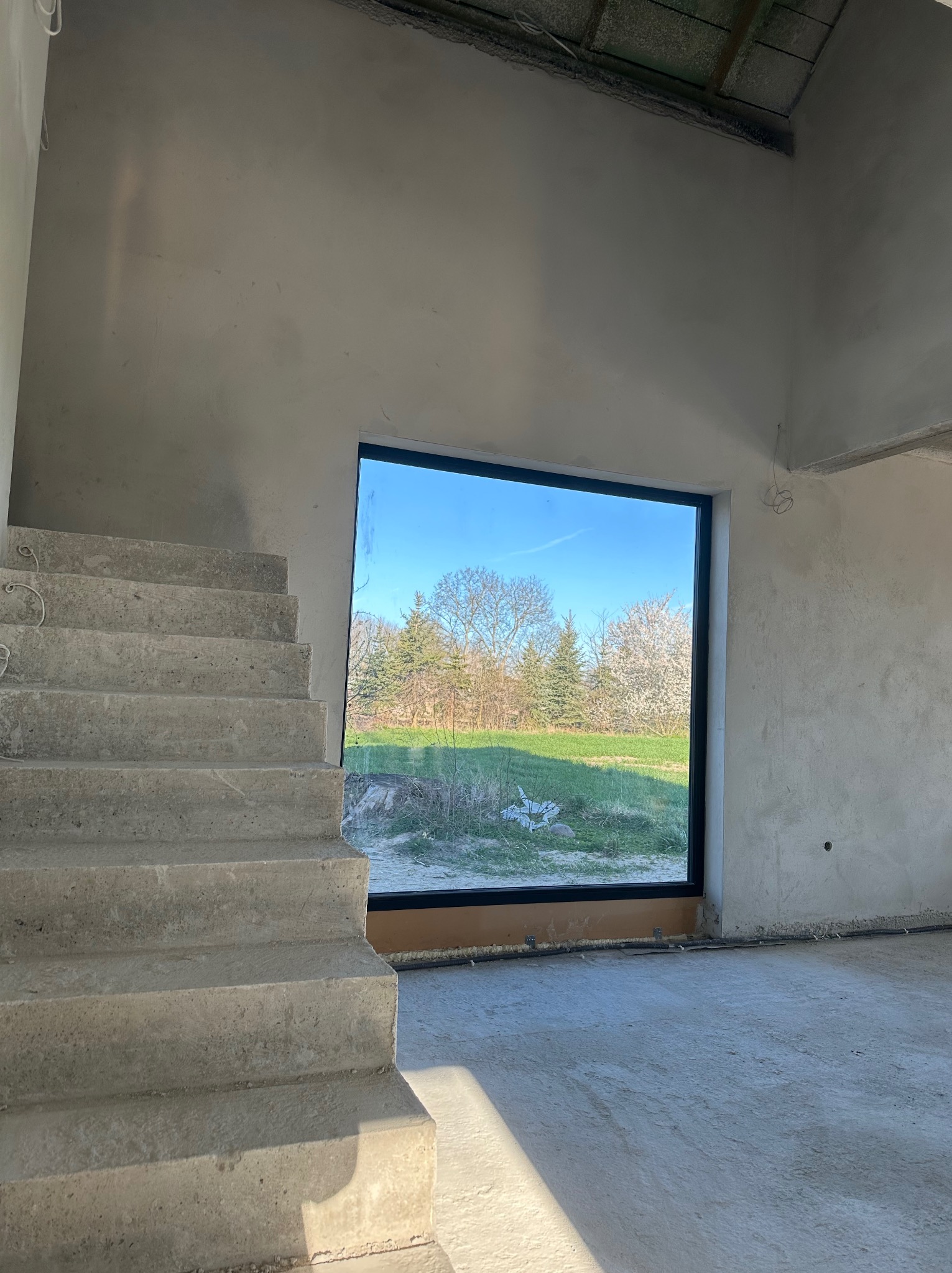 Surowe wnętrze w trakcie prac wykończeniowych: widoczne betonowe schody, ściany pokryte szarym tynkiem oraz duże okno z widokiem na zewnątrz, gdzie widać zieleń i drzewa.