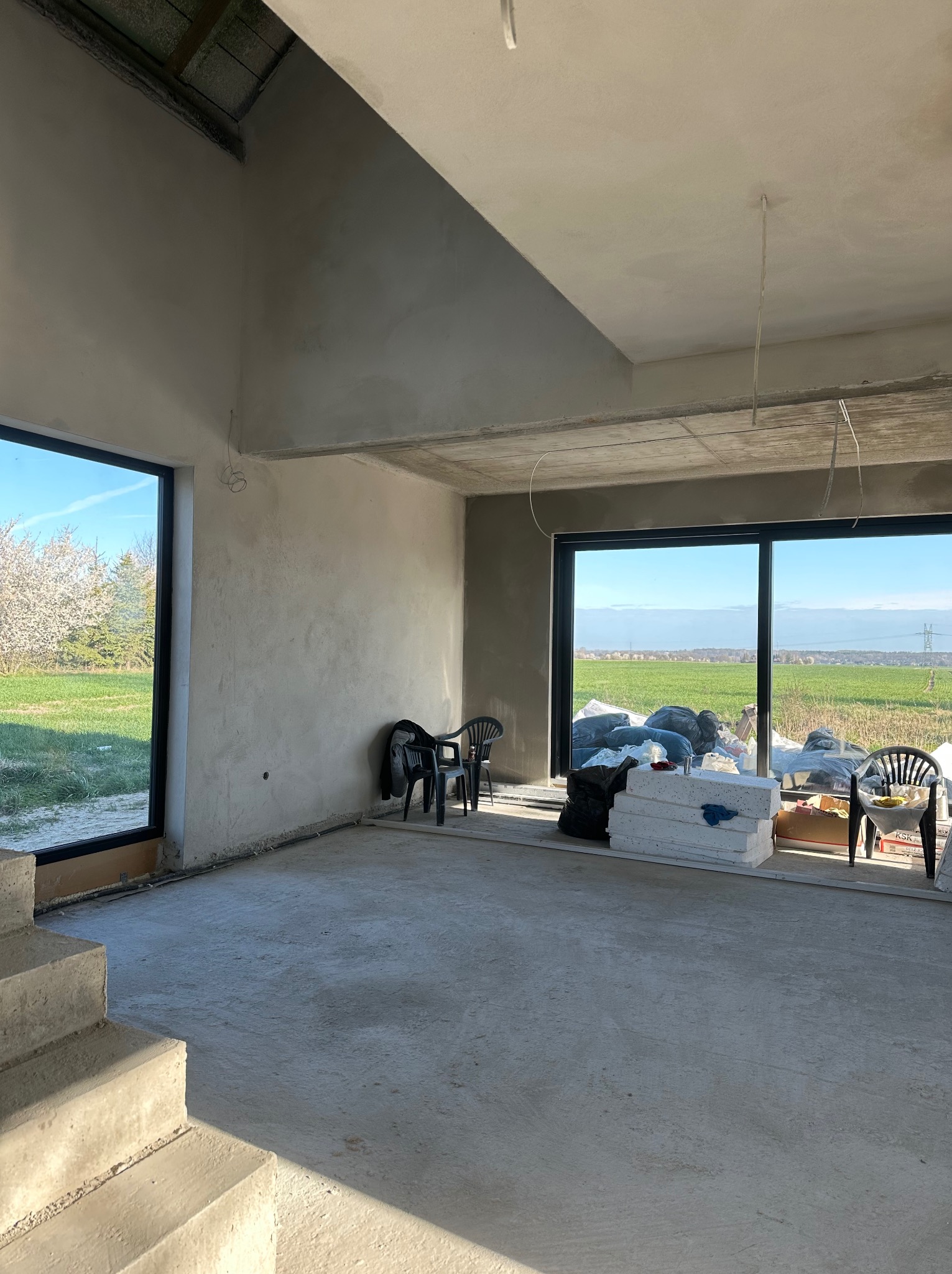 Surowe wnętrze domu w trakcie tynkowania maszynowego; widoczne gładkie, szare ściany, betonowe schody, duże okna z widokiem na zielone pole i drzewa, worki z materiałami budowlanymi i plastikowe...