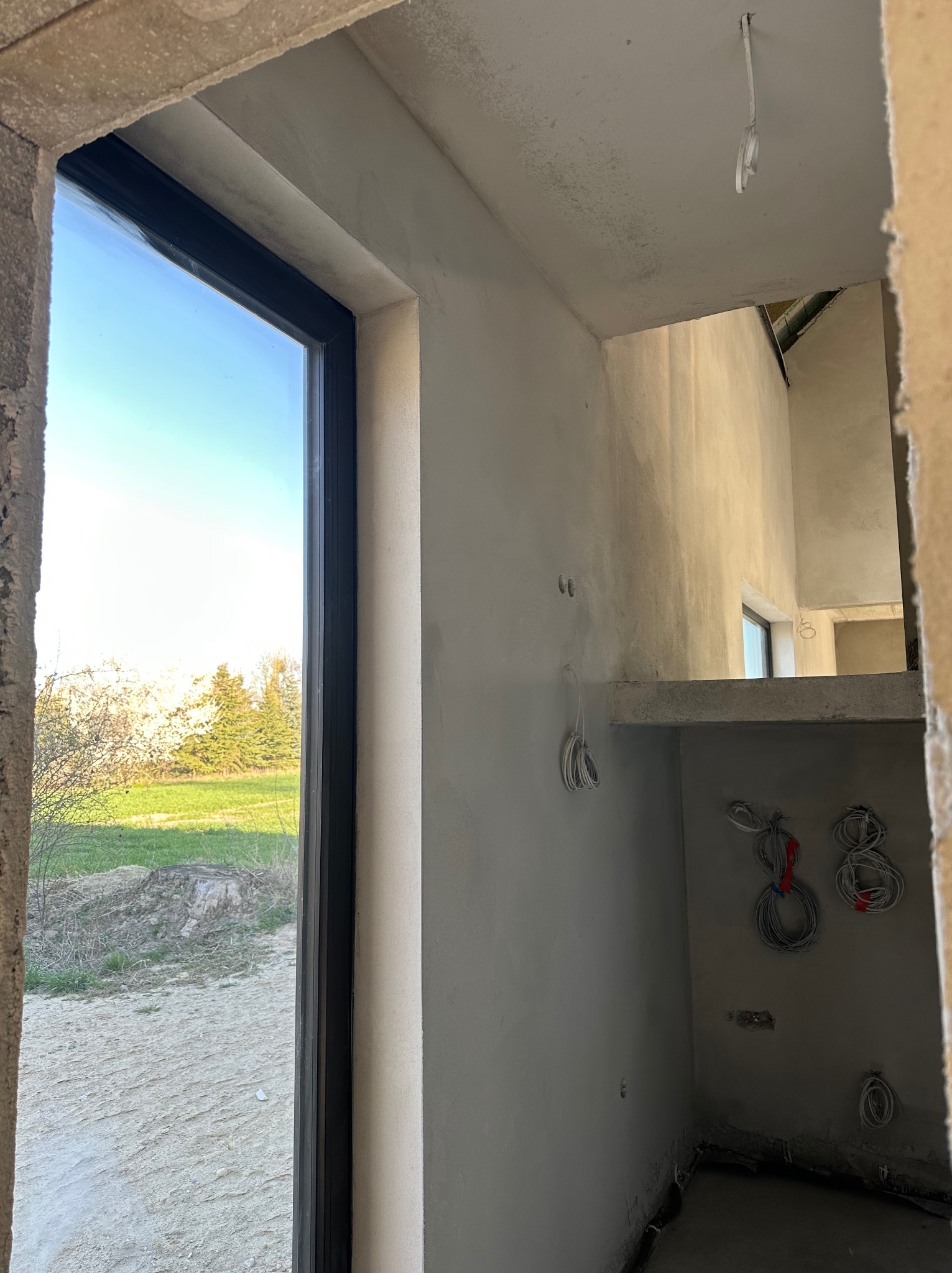 Wnętrze w trakcie prac wykończeniowych; widok przez okno na zewnątrz; ściany pokryte świeżym tynkiem, widoczne przewody elektryczne zwisające ze ściany i sufitu.