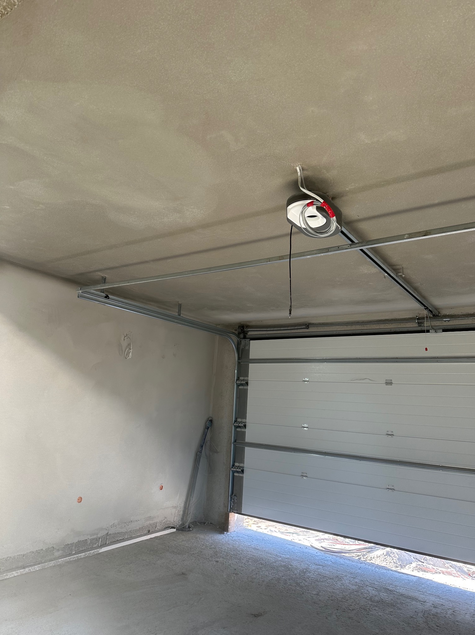 Surowy garaż z świeżo nałożonym tynkiem na ścianach i suficie, widoczny mechanizm otwierania bramy garażowej, brama częściowo otwarta.