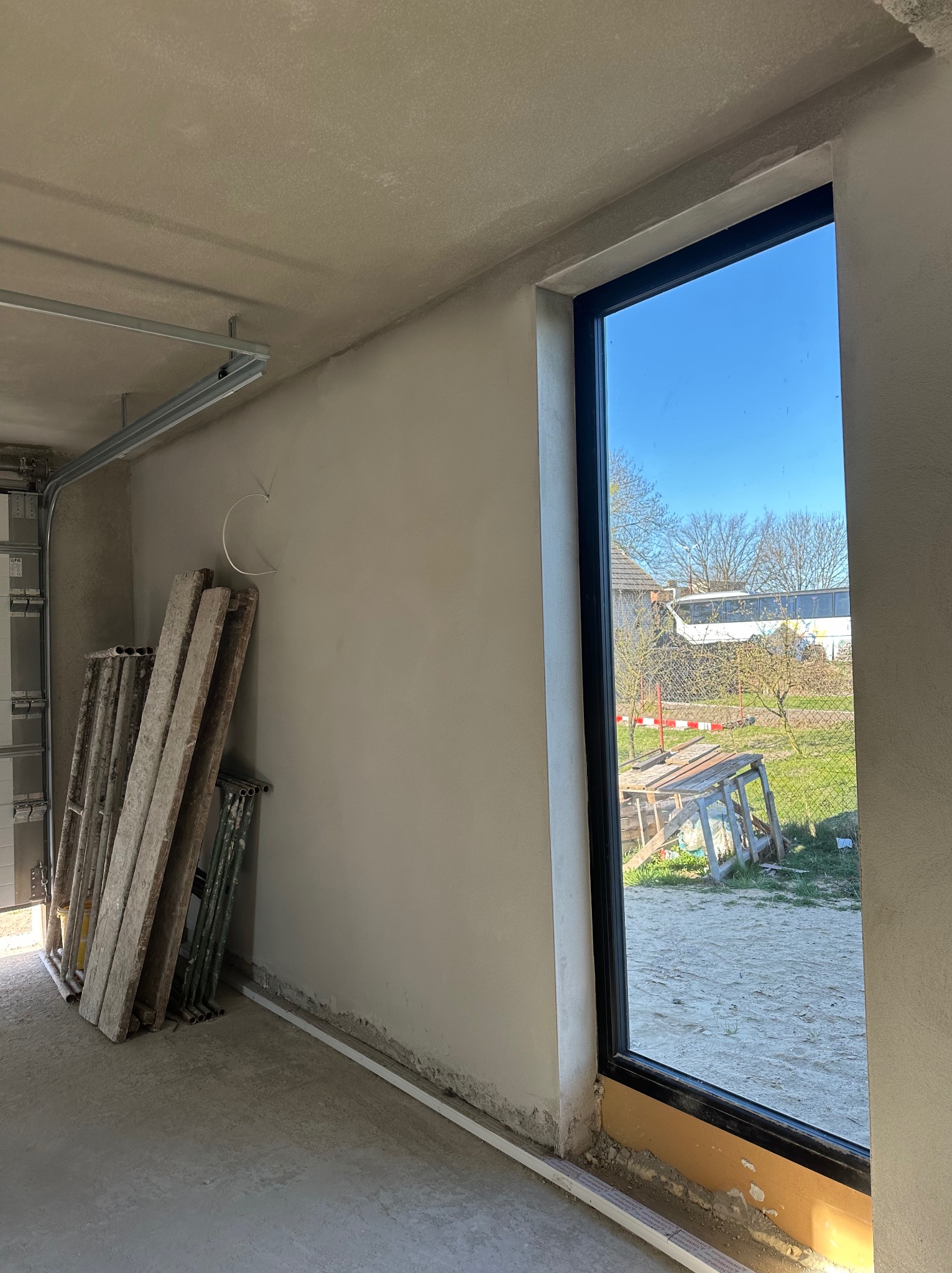 Wnętrze w trakcie prac wykończeniowych: ściana po nałożeniu tynku, widoczne rusztowania oparte o ścianę, okno z widokiem na ogród i niebo.