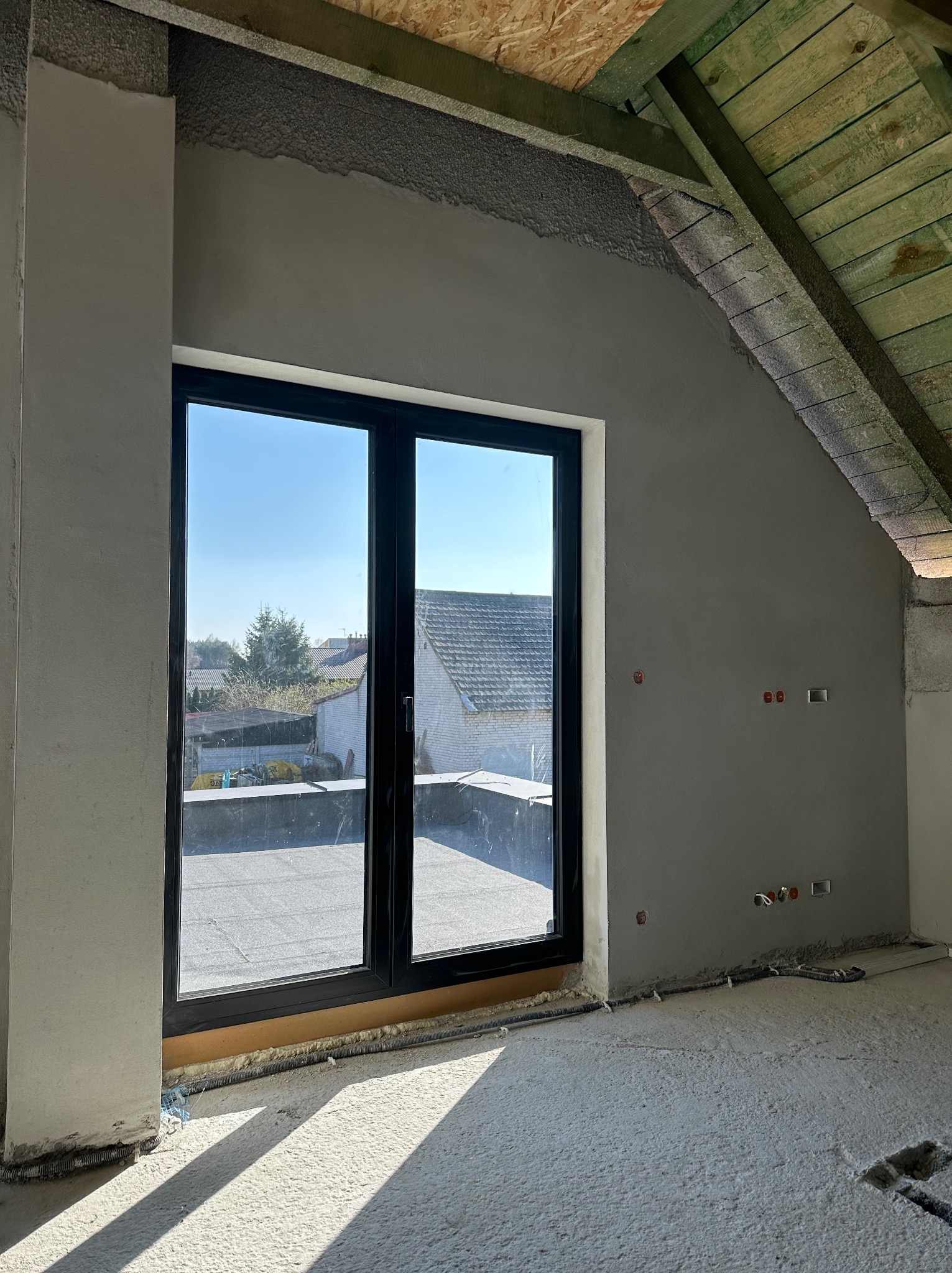 Wnętrze poddasza w trakcie prac wykończeniowych; ściany pokryte szarą, świeżo nałożoną warstwą, widoczne puszki elektryczne, czarne okno z widokiem na dachy domów i drzewa, surowe drewniane krokwie...